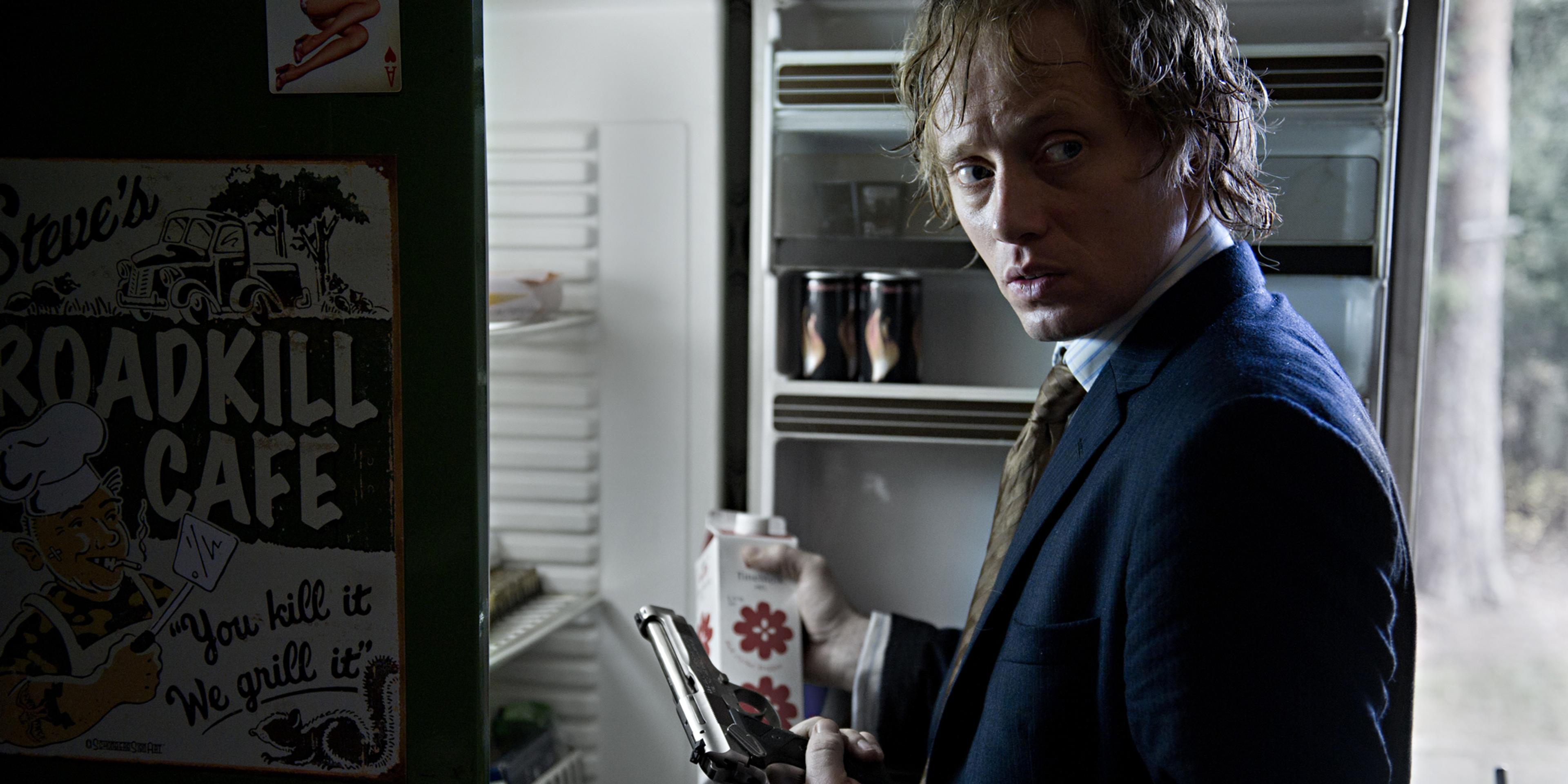 En mann står foran et kjøleskap med en pistol i den ene hånden og en melkekartong i den andre