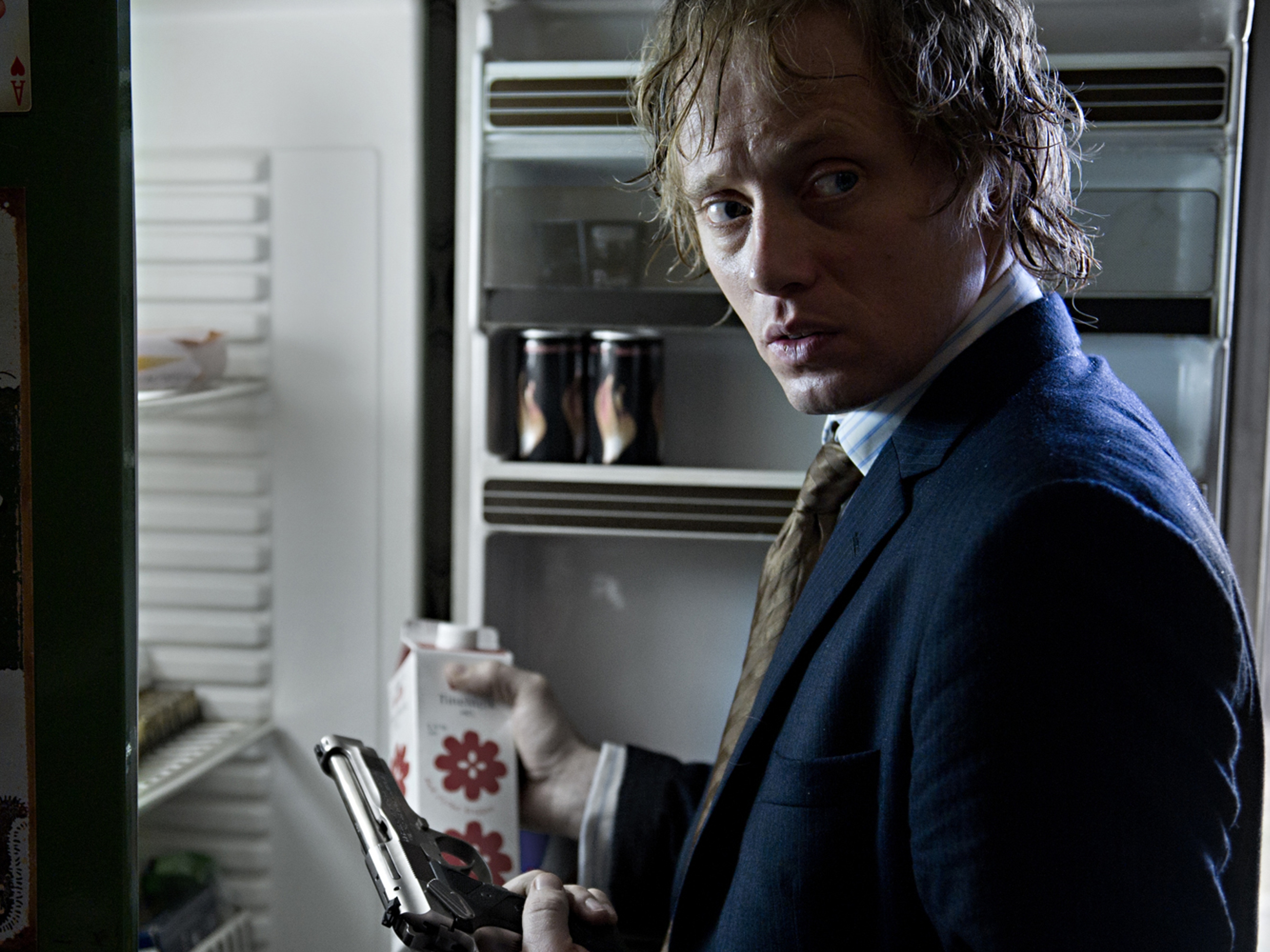 En mann står foran et kjøleskap med en pistol i den ene hånden og en melkekartong i den andre