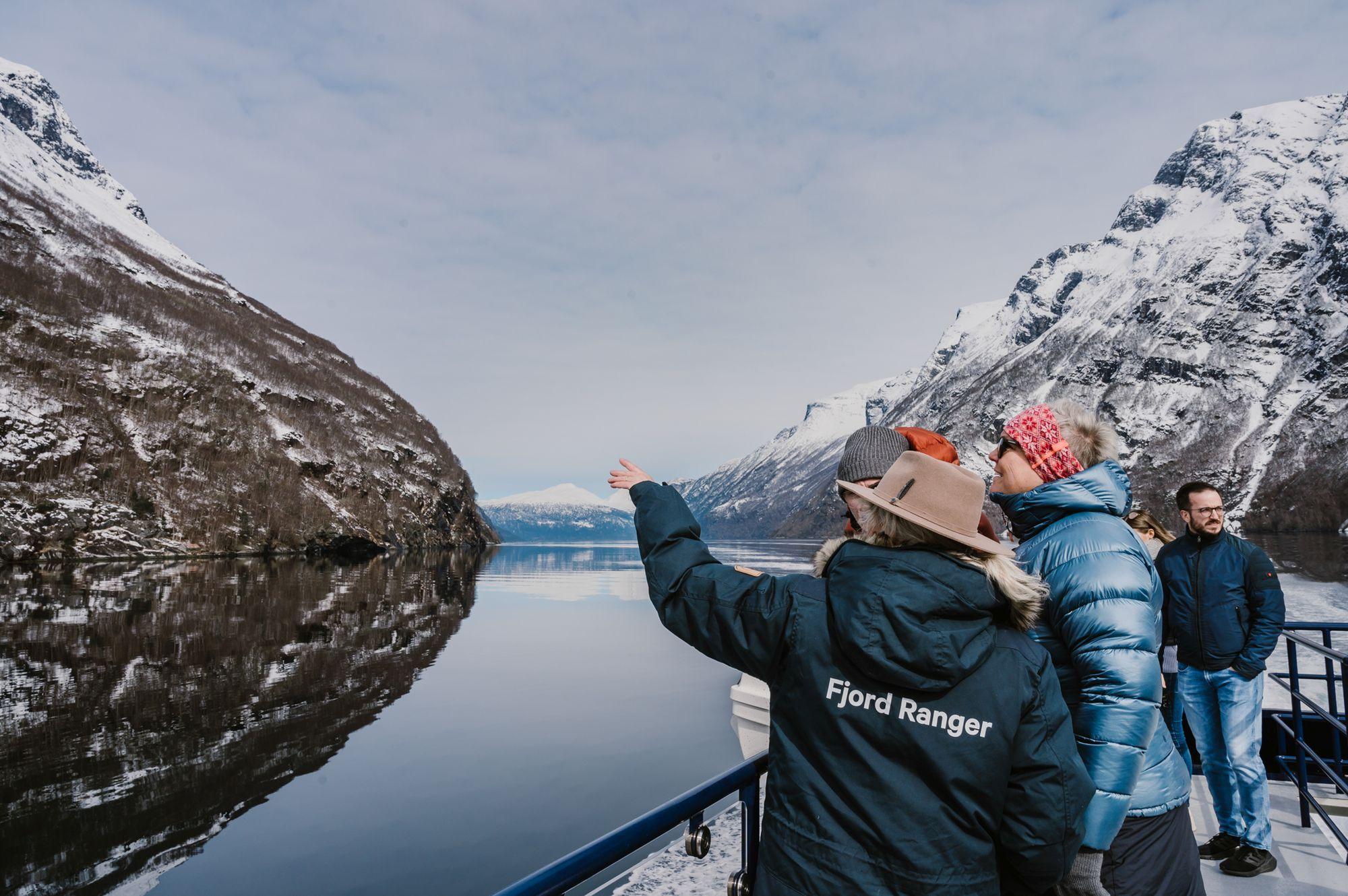 Tourists on a boat deck viewing snow-capped mountains reflected in calm fjord waters in Geirangerfjord