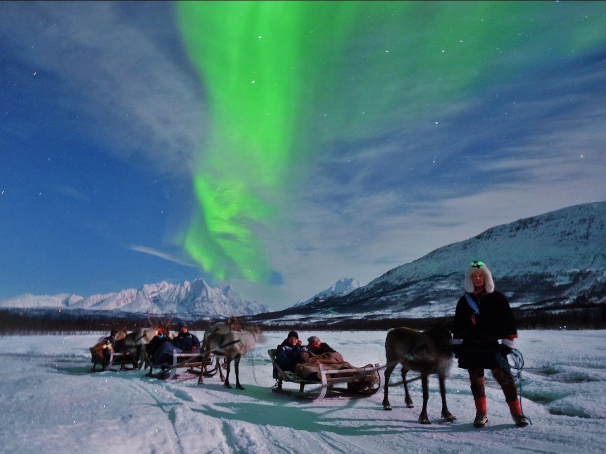 Aurora over snowy landscape with travelers in reindeer sleds