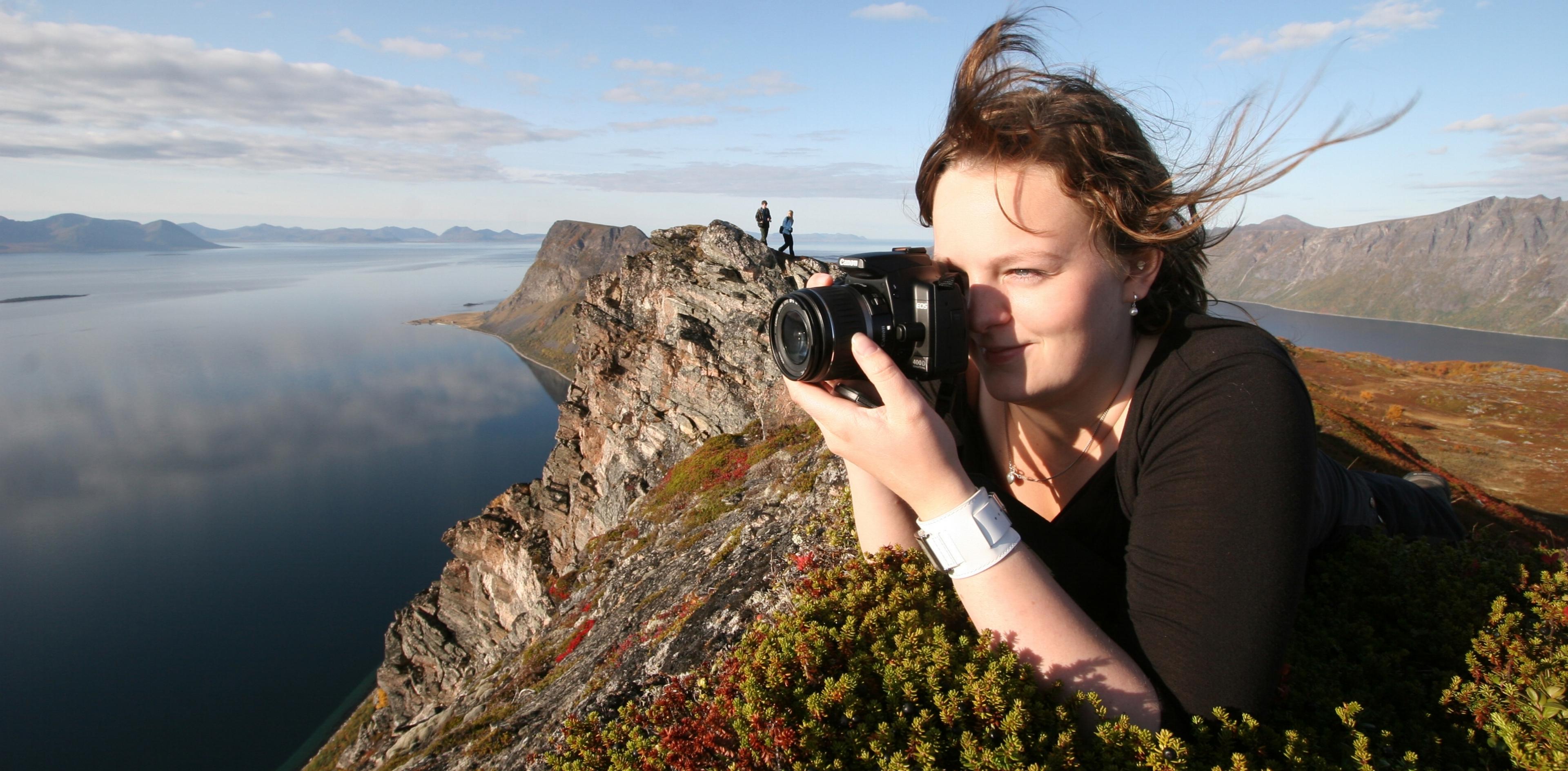 En jente ligger i lyngen på toppen av et fjell og tar bilde av horisonten