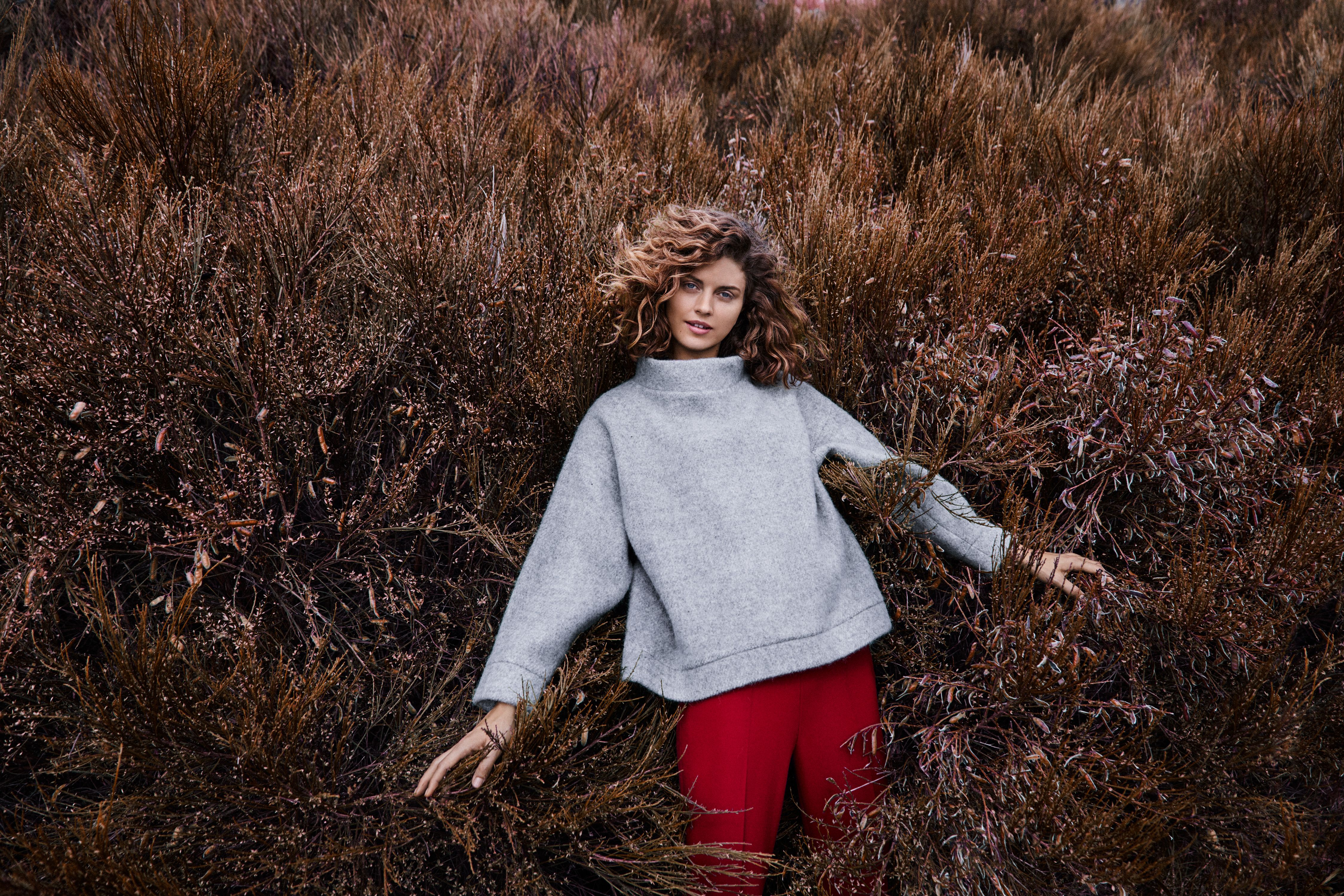 A girl wearing a woollen sweater, standing in a field with bushes