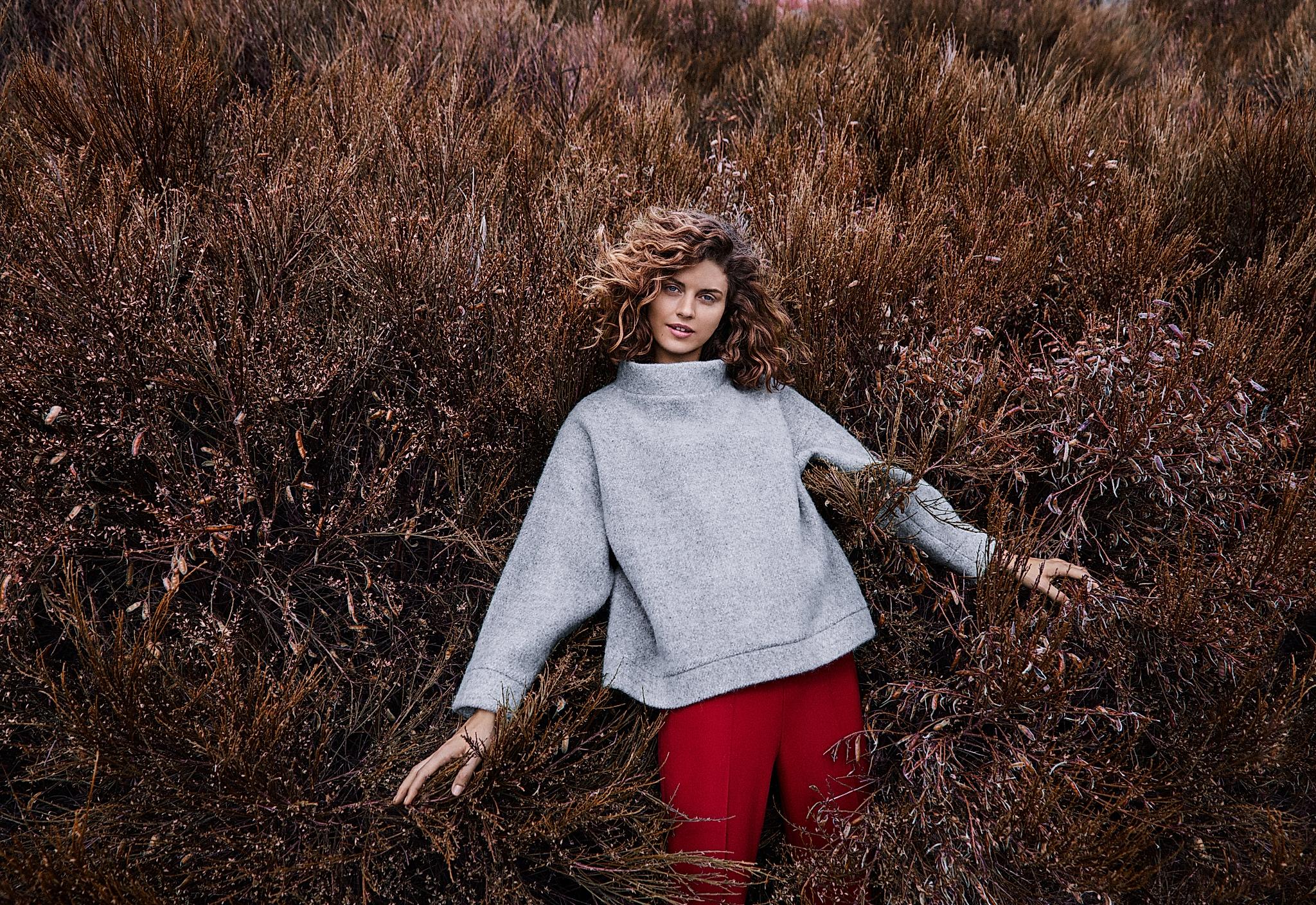 A girl wearing a woollen sweater, standing in a field with bushes