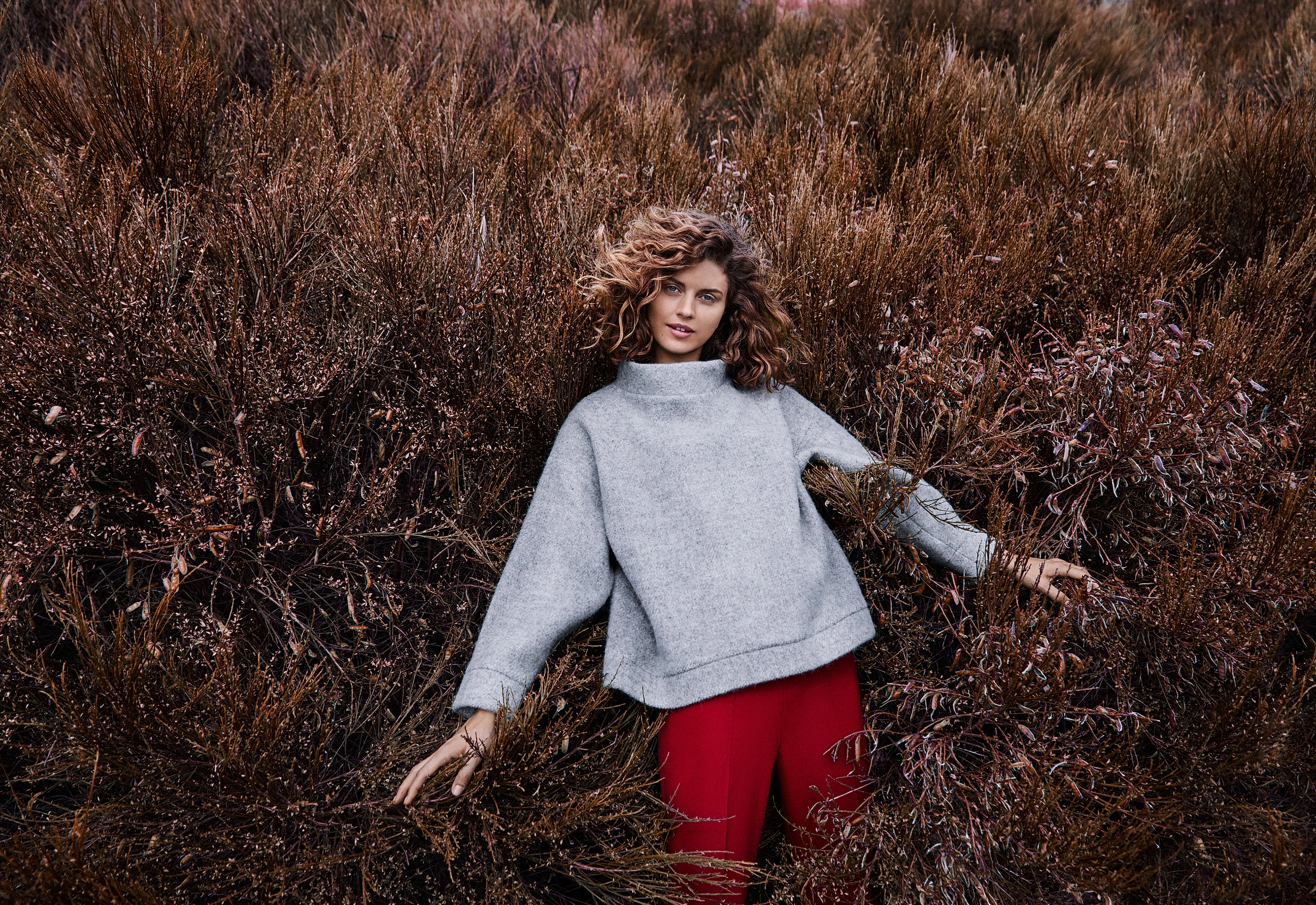 A girl wearing a woollen sweater, standing in a field with bushes