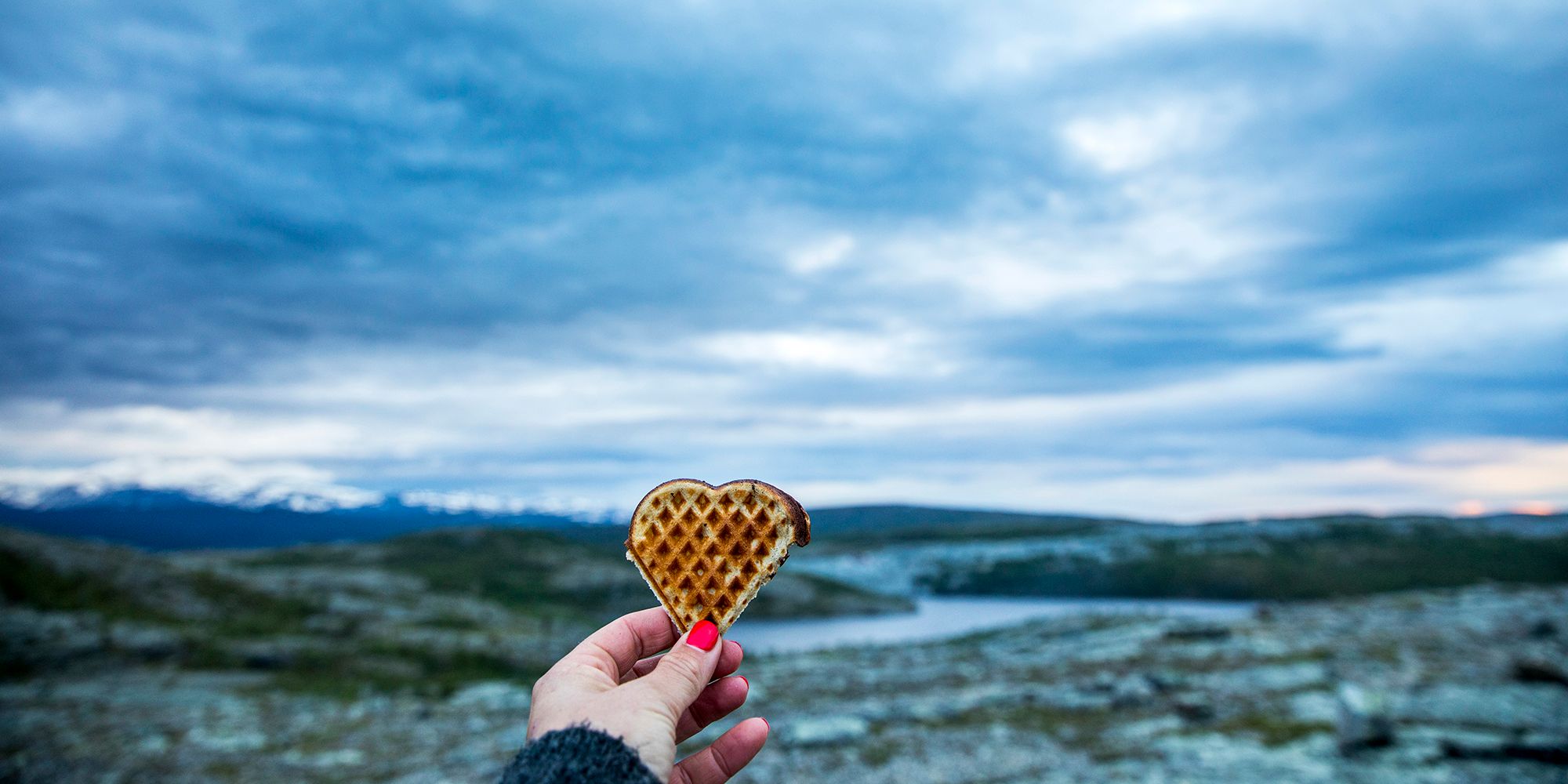 A hand holding up a heart-shaped waffle with beautiful scenery in the background