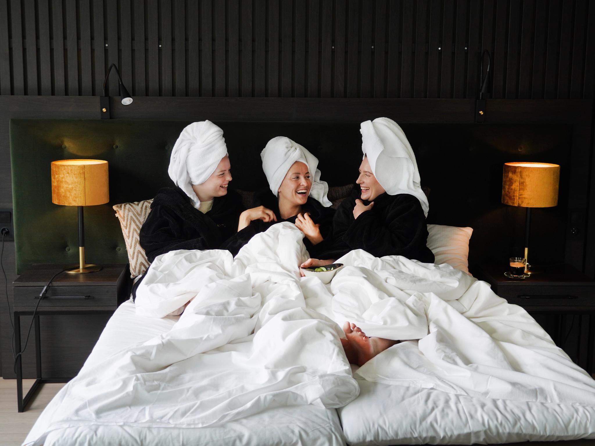Tre damer som smiler i en hotellseng.