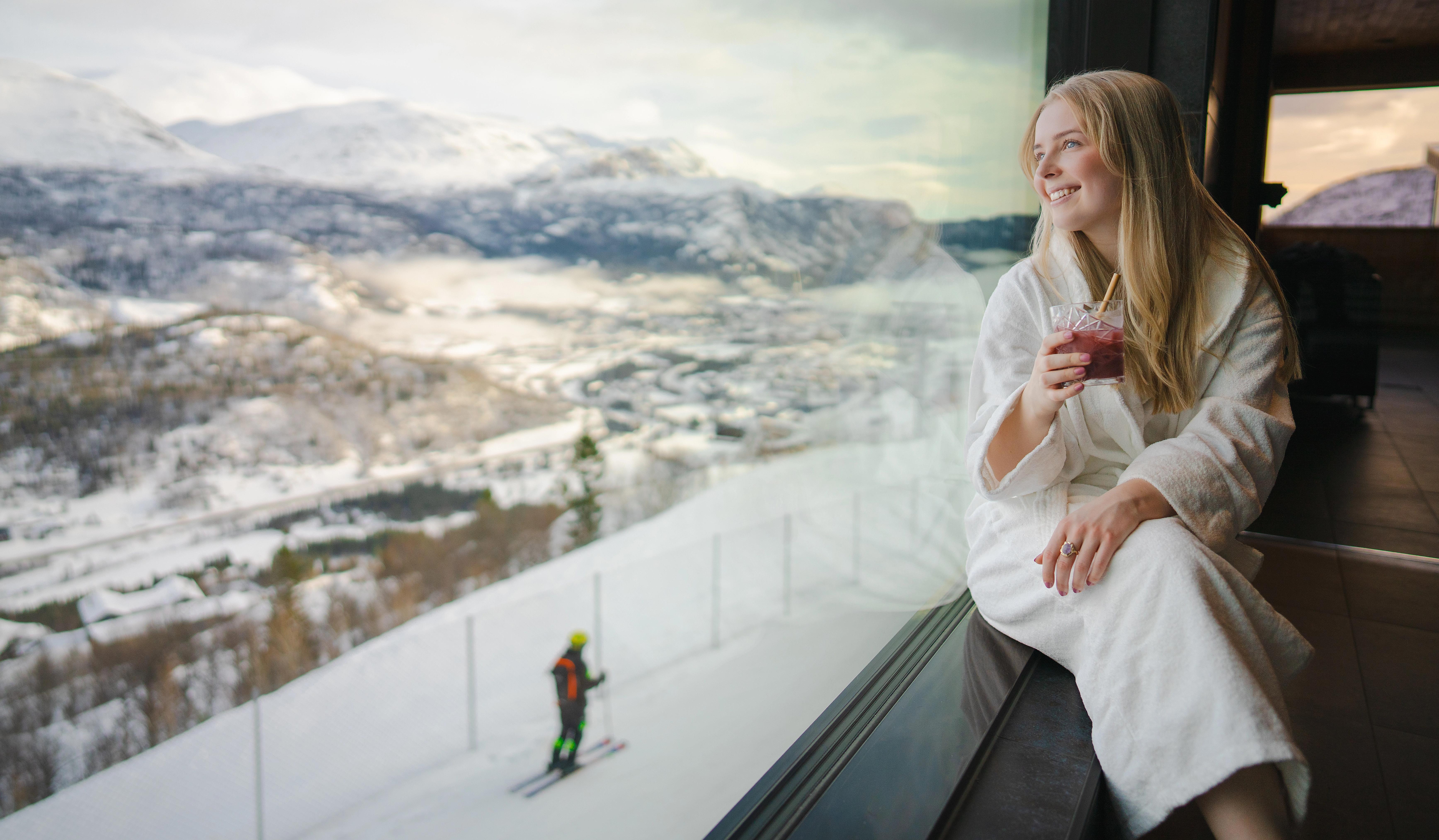 En jente slapper av inne på spaet på Skarsnuten, mens en skiløper suser nedover bakkene i bakgrunnen