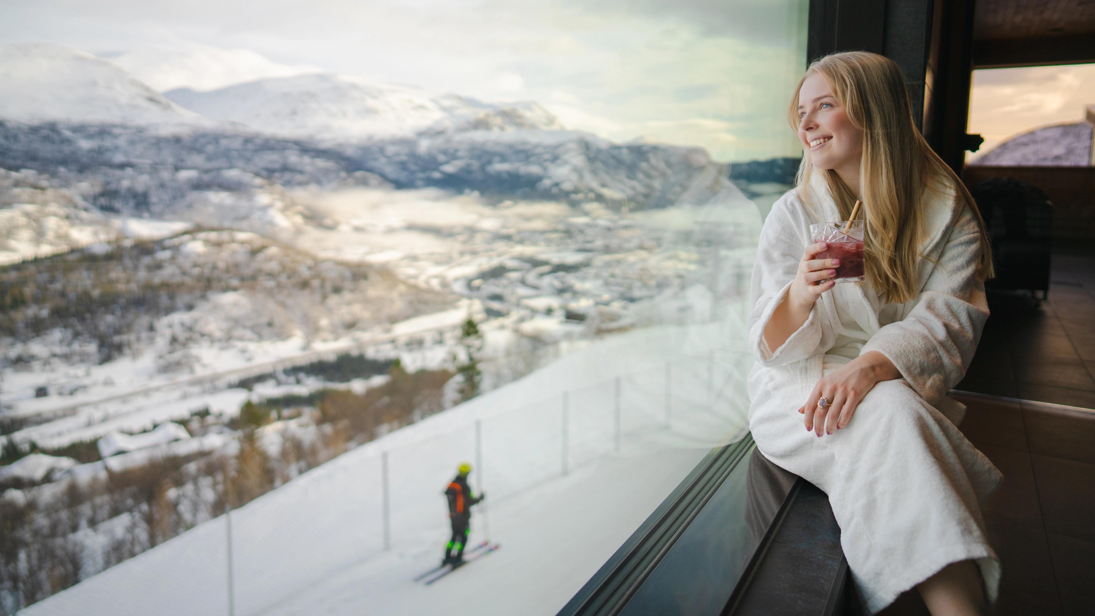 A girl relaxes inside the spa at Skarsnuten, while a skier glides down the slopes in the background