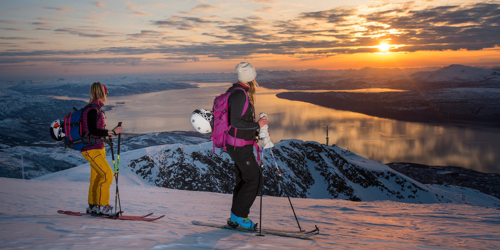 Two women with skis enjoying the view of mountains and fjord from the top of a mountain in Narvik, Northern Norway