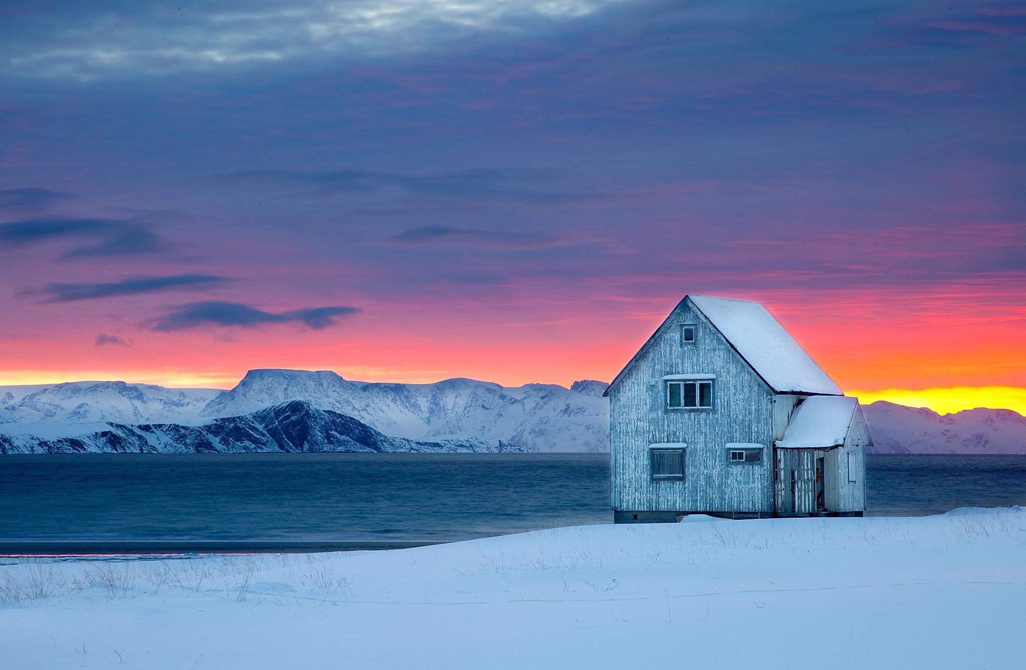 A wooden house next to the water on a snow-covered landscape with sunset colors in the background at Hasvik in Finnmark, Northern Norway