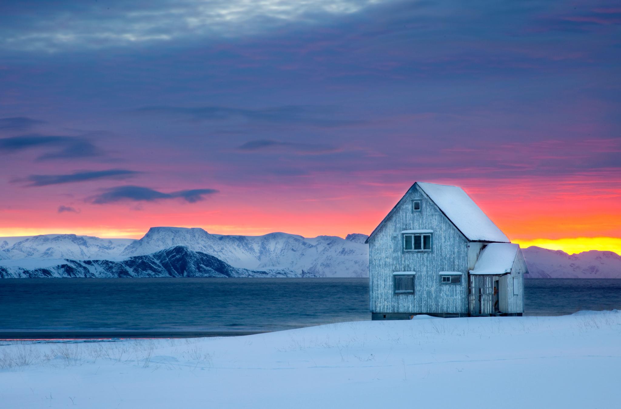 A wooden house next to the water on a snow-covered landscape with sunset colors in the background at Hasvik in Finnmark, Northern Norway