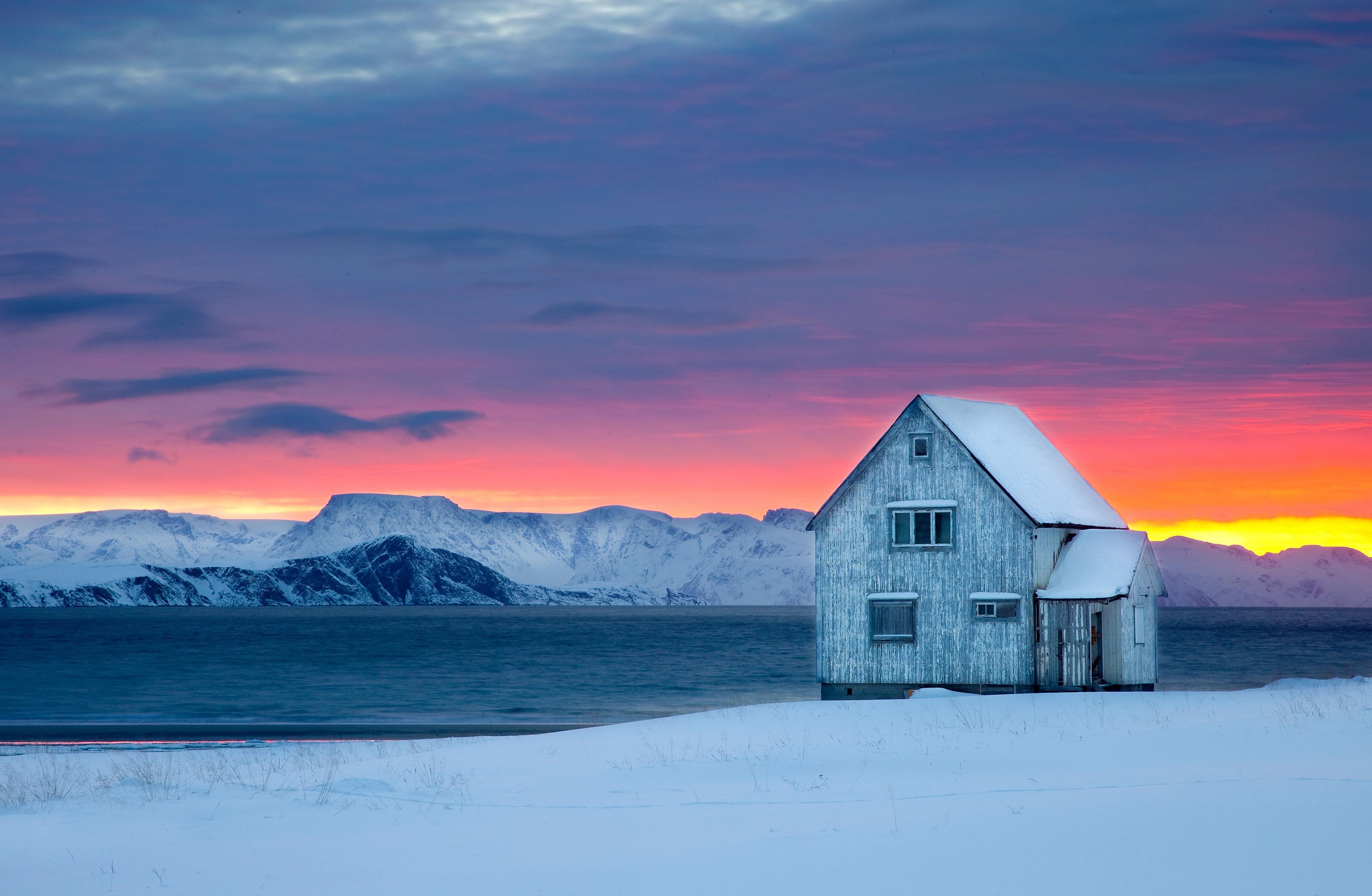 A wooden house next to the water on a snow-covered landscape with sunset colors in the background at Hasvik in Finnmark, Northern Norway