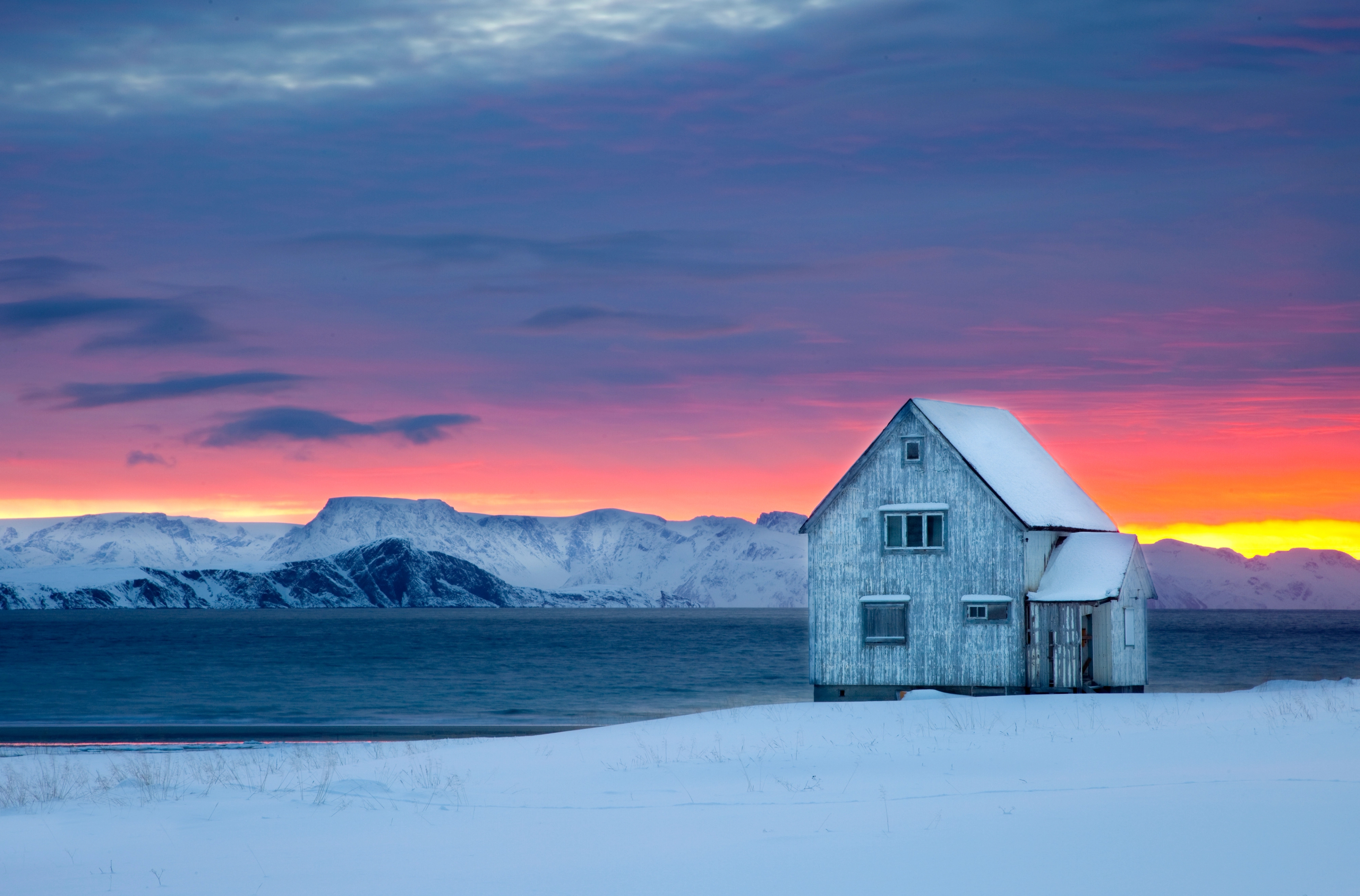 A wooden house next to the water on a snow-covered landscape with sunset colors in the background at Hasvik in Finnmark, Northern Norway