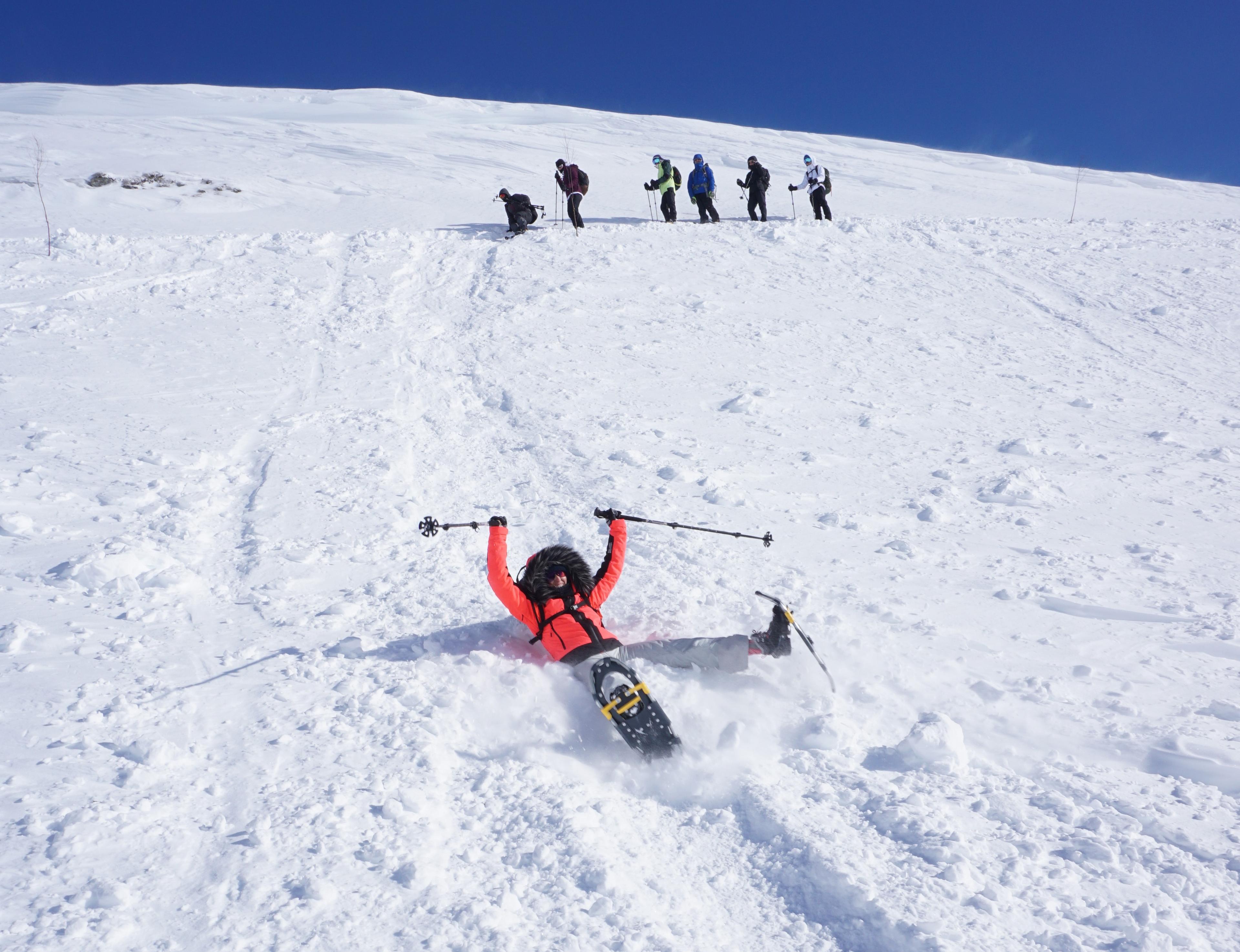 A group is snowshoeing to Trolltunga in Fjord Norway in winter, and one woman is sliding down the hill on her butt, smiling.