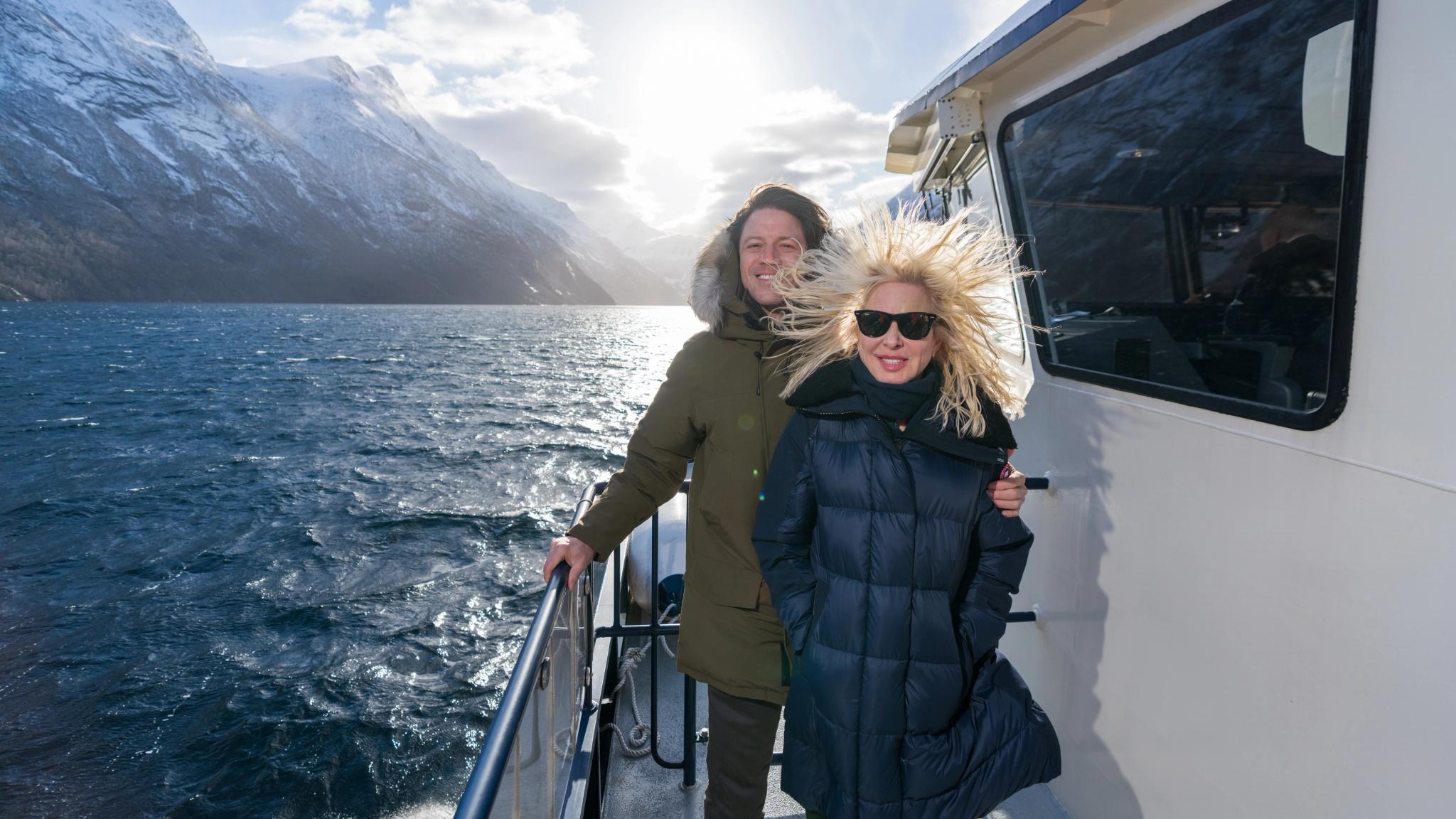 Mann og kvinne i vinden på taket av Fjordrangers cruisebåt til Geiranger om vinteren