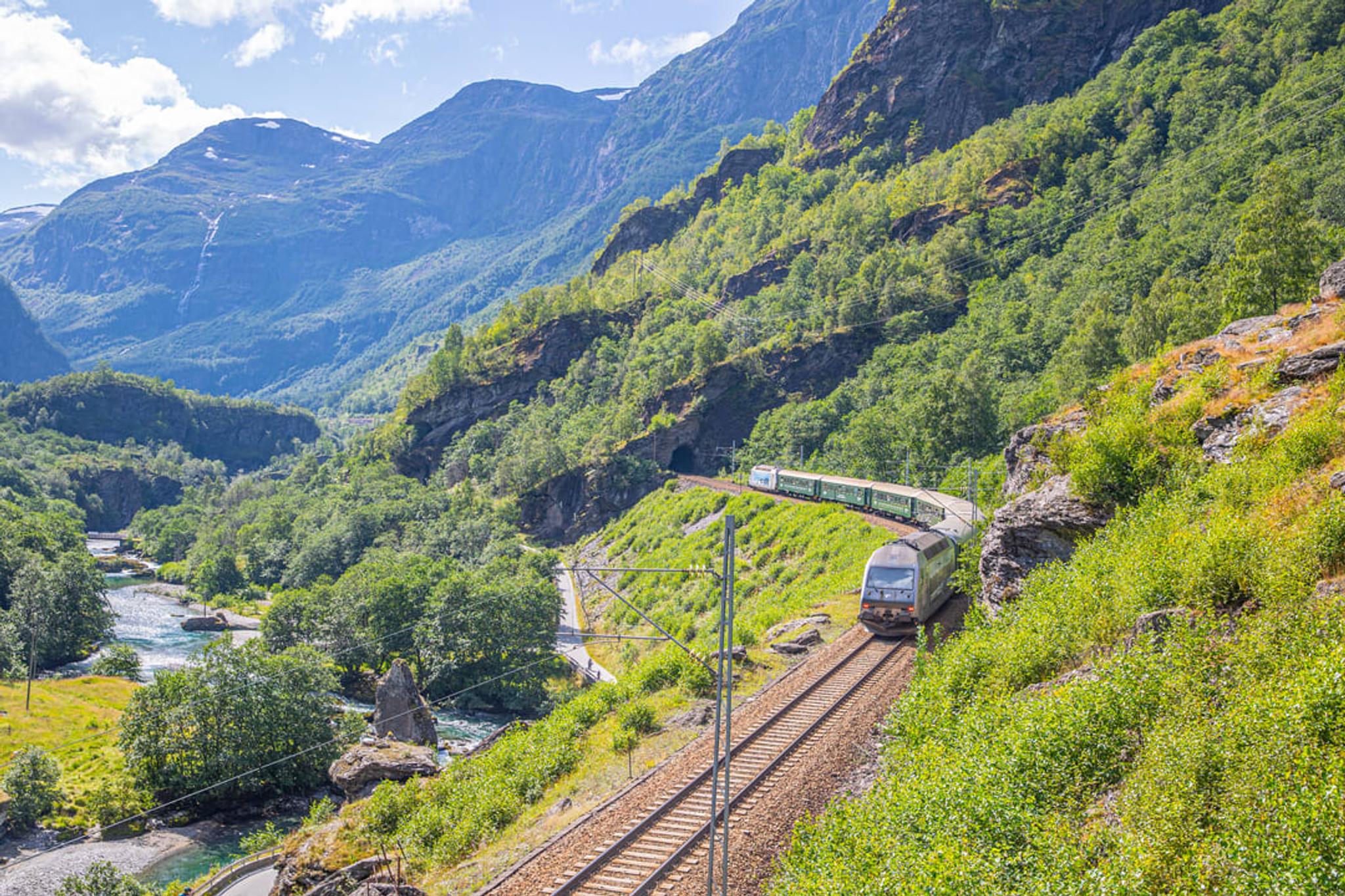 The Flåm Railway train travelling along a steep hillside, with a river valley and mountain peaks in the background.