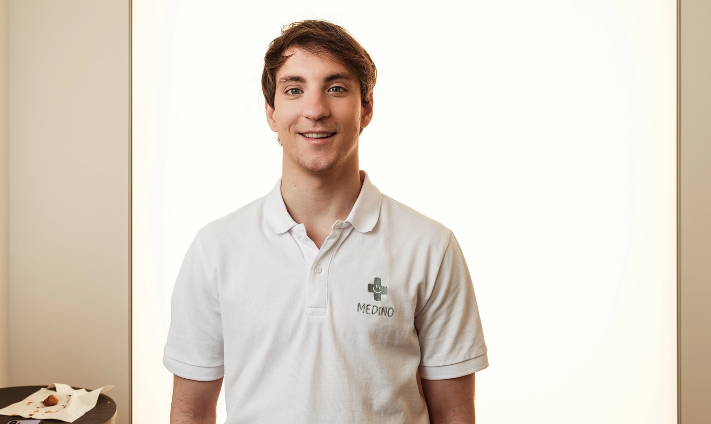 Male employee of ST Healthcare in a white polo with a small green cross on the left chest and dark pants, smiling