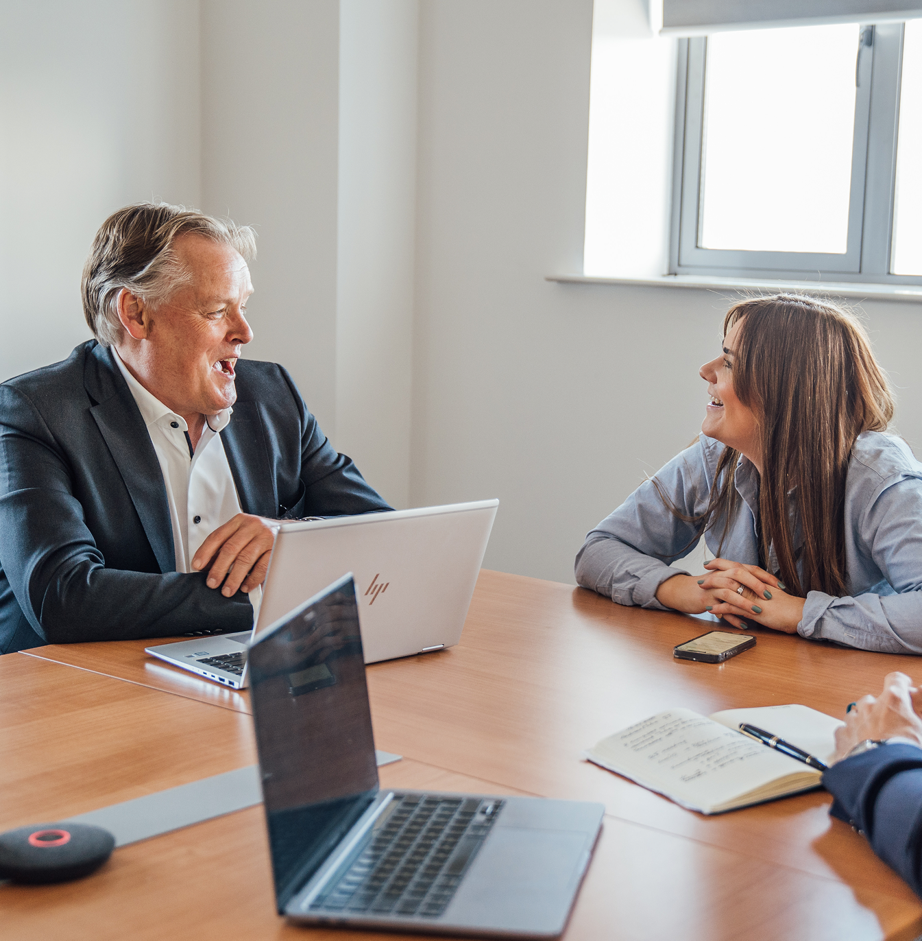 Two colleagues smiling and talking across a conference table with laptops and a notepad during an office meeting