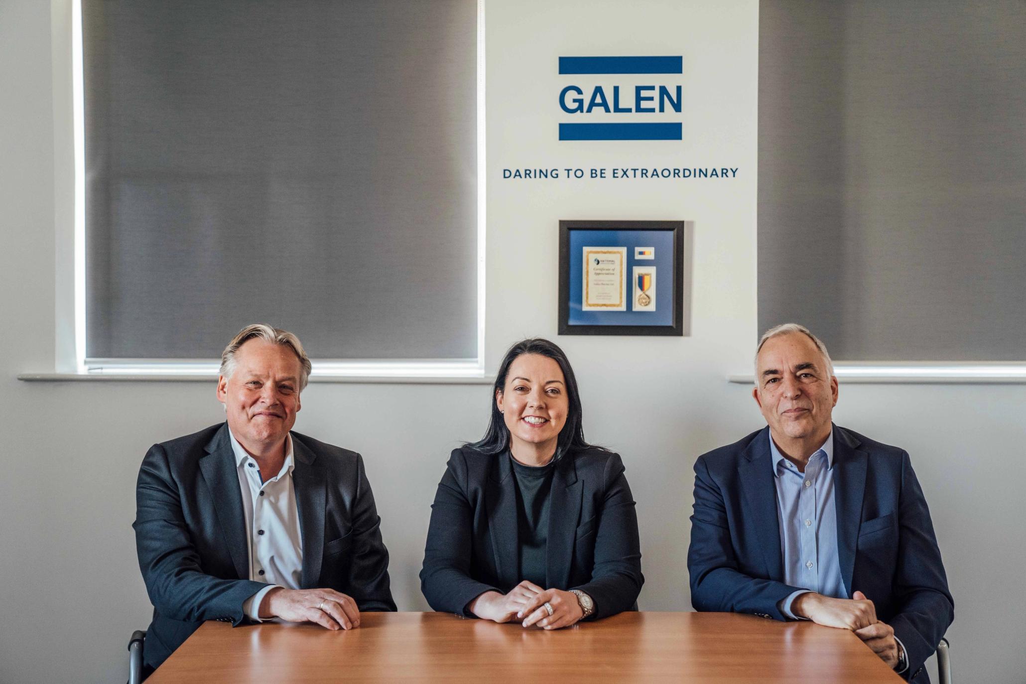 Drei Geschäftsleute sitzen lächelnd an einem Konferenztisch vor einem Galen-Firmenlogo