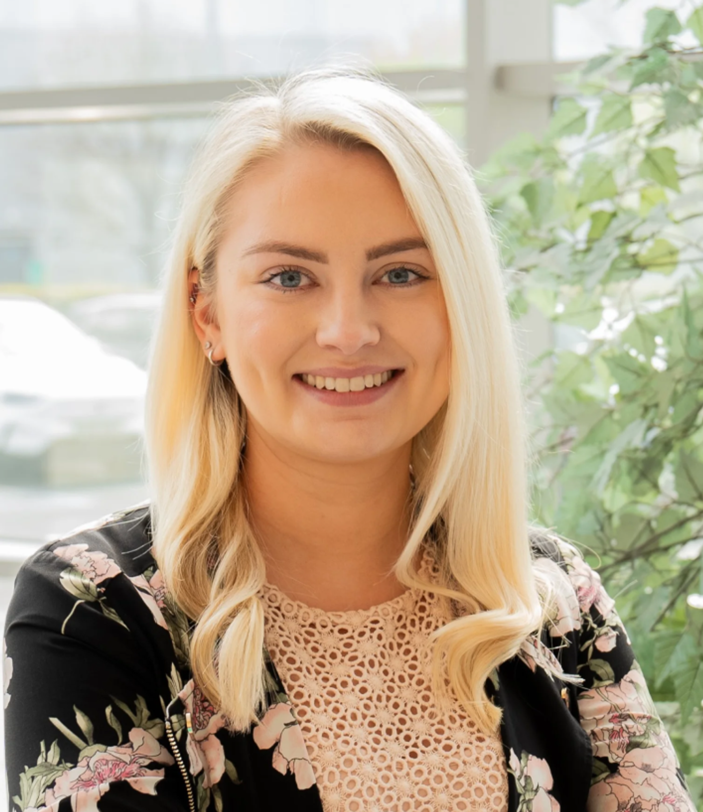 Aimee Lappin smiling in a floral top with lace detail, photographed indoors near a window with greenery