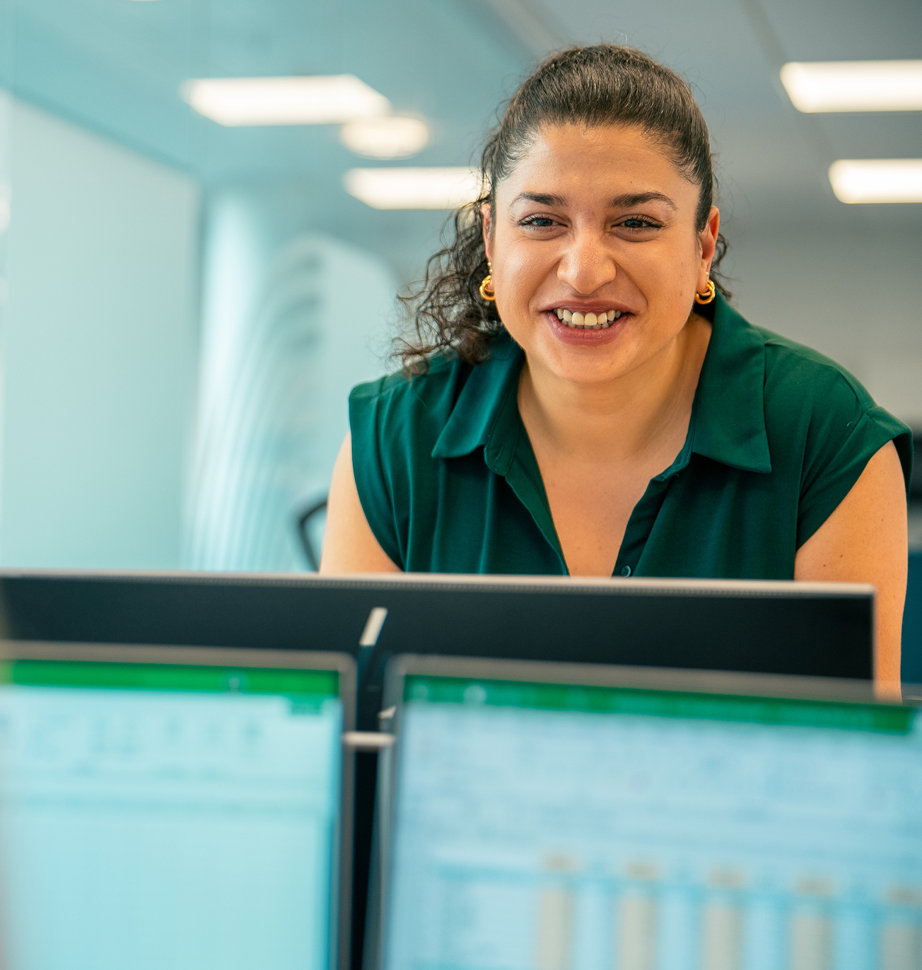 Smiling professional in a green top leaning over a desk with dual computer monitors in a bright office