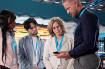 Four conference attendees with lanyards gathered around a person viewing content on a smartphone