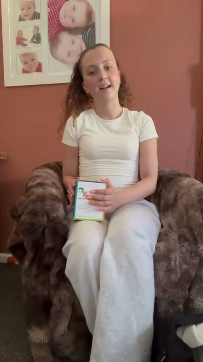 Young woman sitting in an armchair holding a Microtabs pack, talking to camera about travelling with PKU