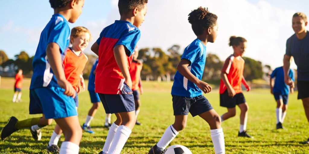 Kinder spielen mit einem Trainer Fußball auf einem Rasenplatz.