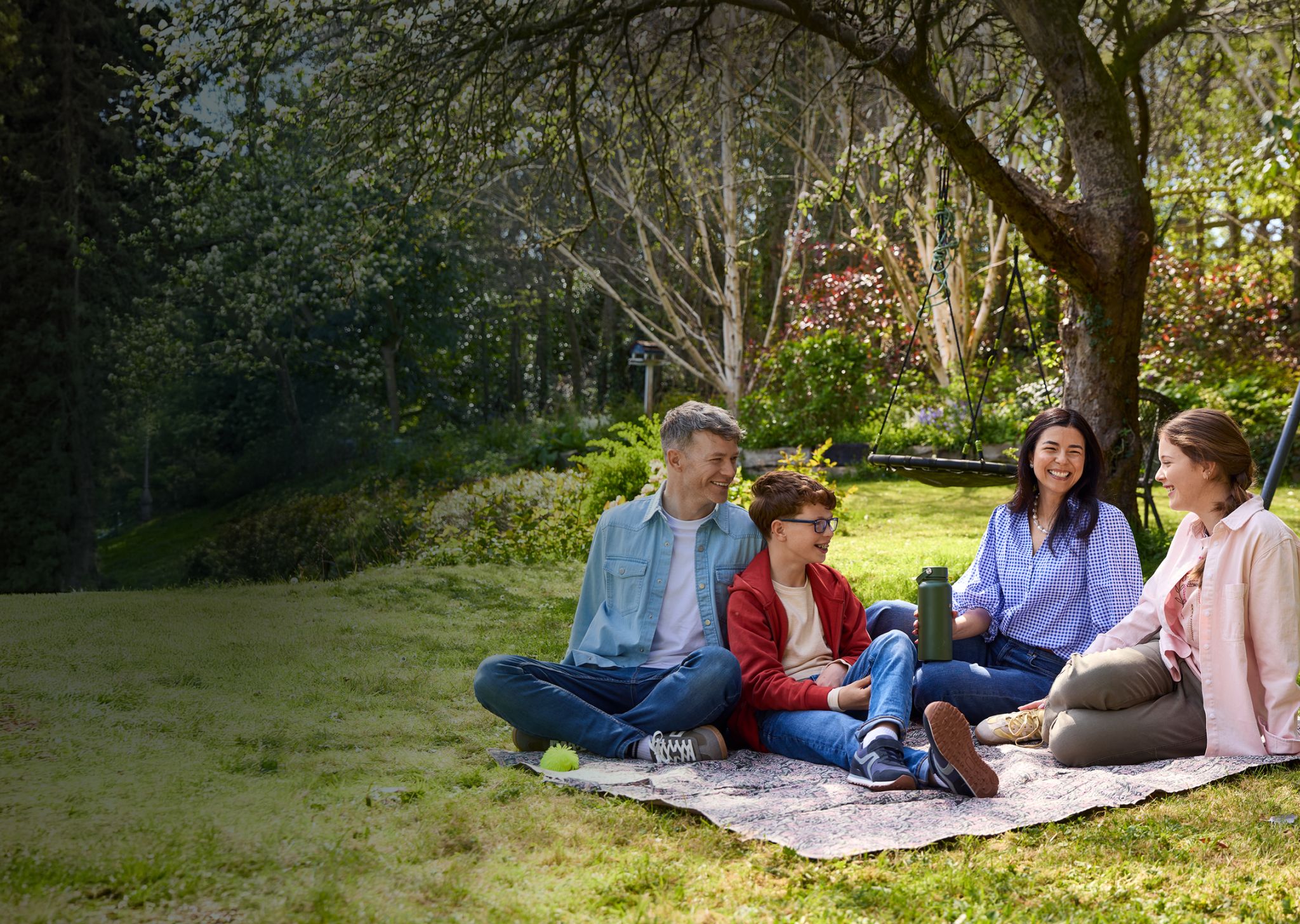 Family relaxing on a picnic blanket under a tree in a sunny, green park