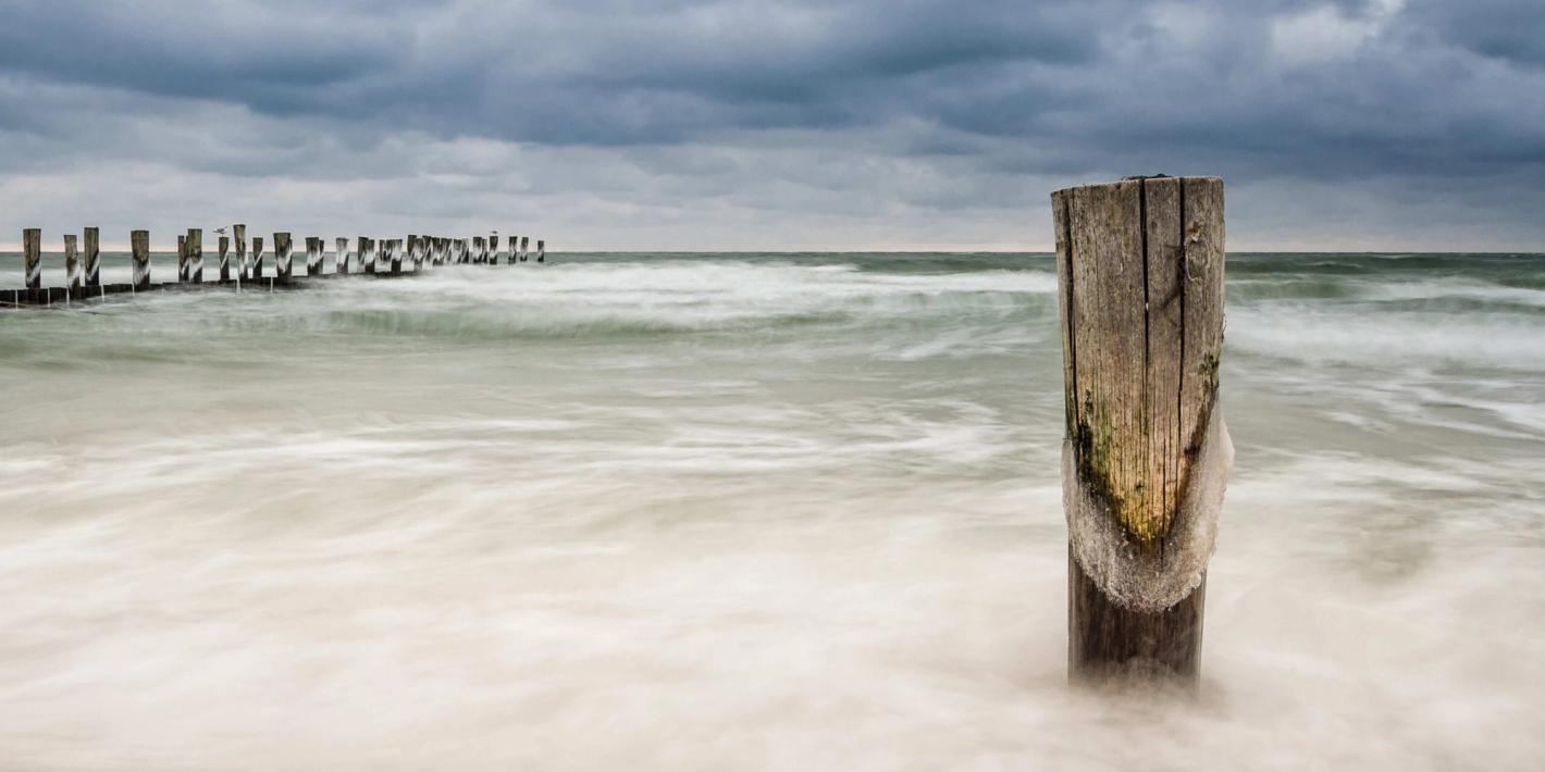 Stürmische See mit Holzpfahl im Vordergrund und Wellen unter dunklen Gewitterwolken