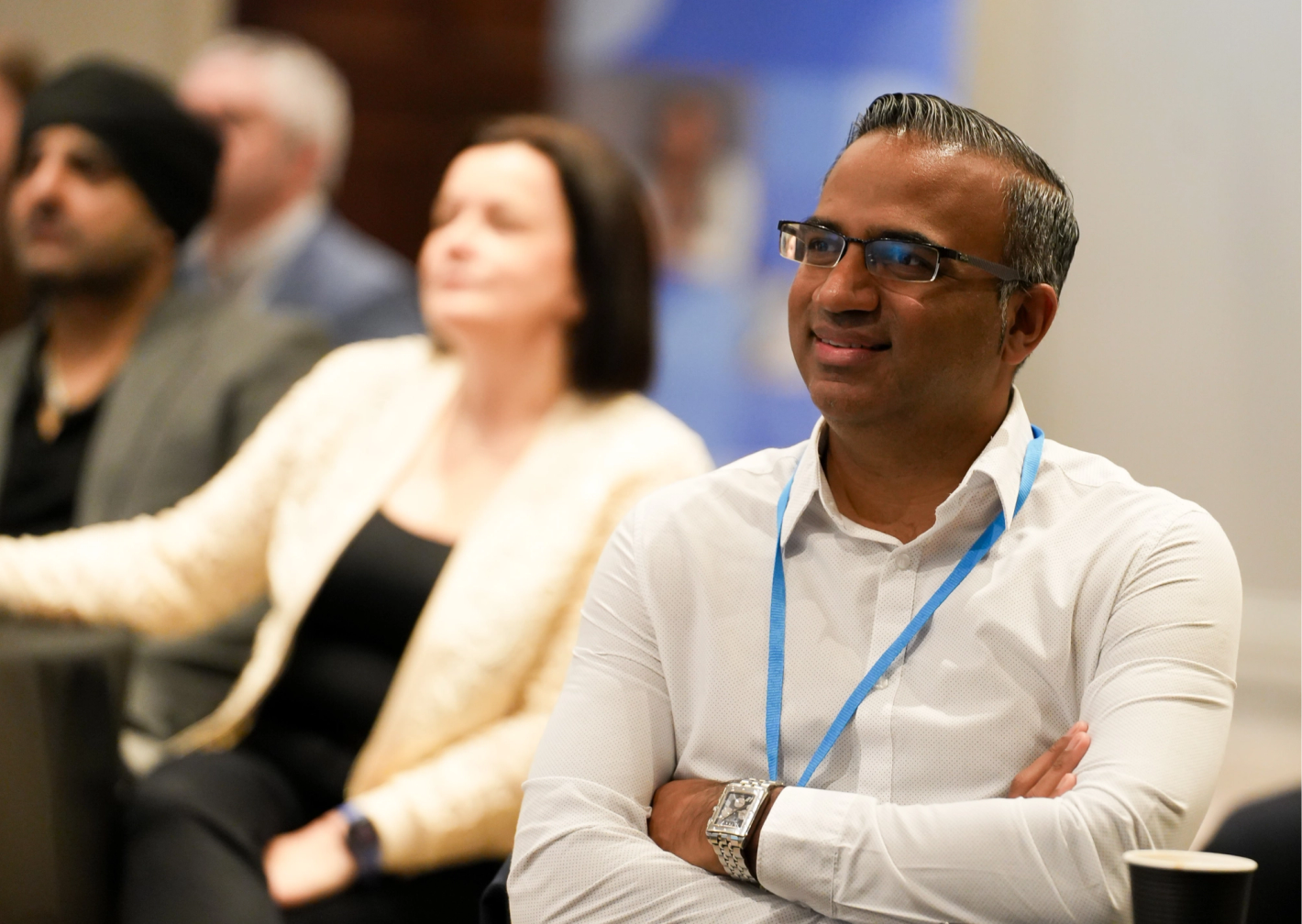 Smiling attendee with glasses and blue lanyard sitting with arms crossed at a professional conference event