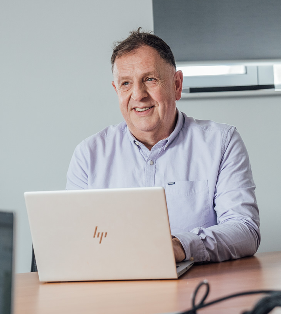 Smiling middle-aged man in a light blue shirt sitting at a desk with an open laptop in an office setting