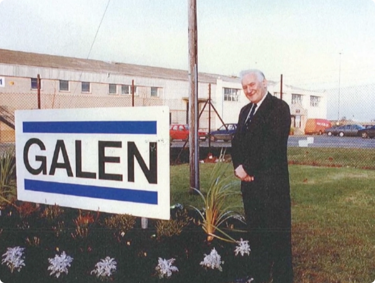Man standing beside a Galen company sign outside an industrial facility in a vintage photograph