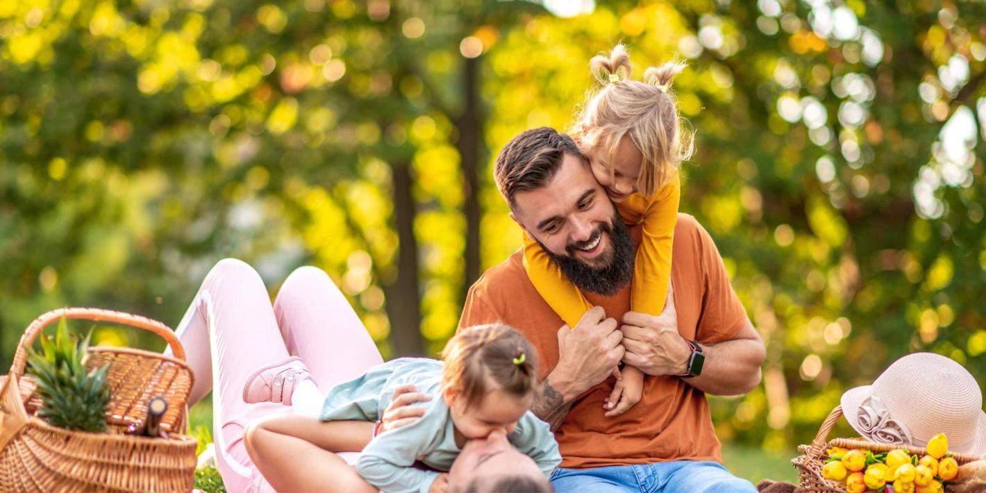 Glückliche Familie beim Picknick im Park, Vater spielt lachend mit zwei kleinen Kindern auf der Wiese
