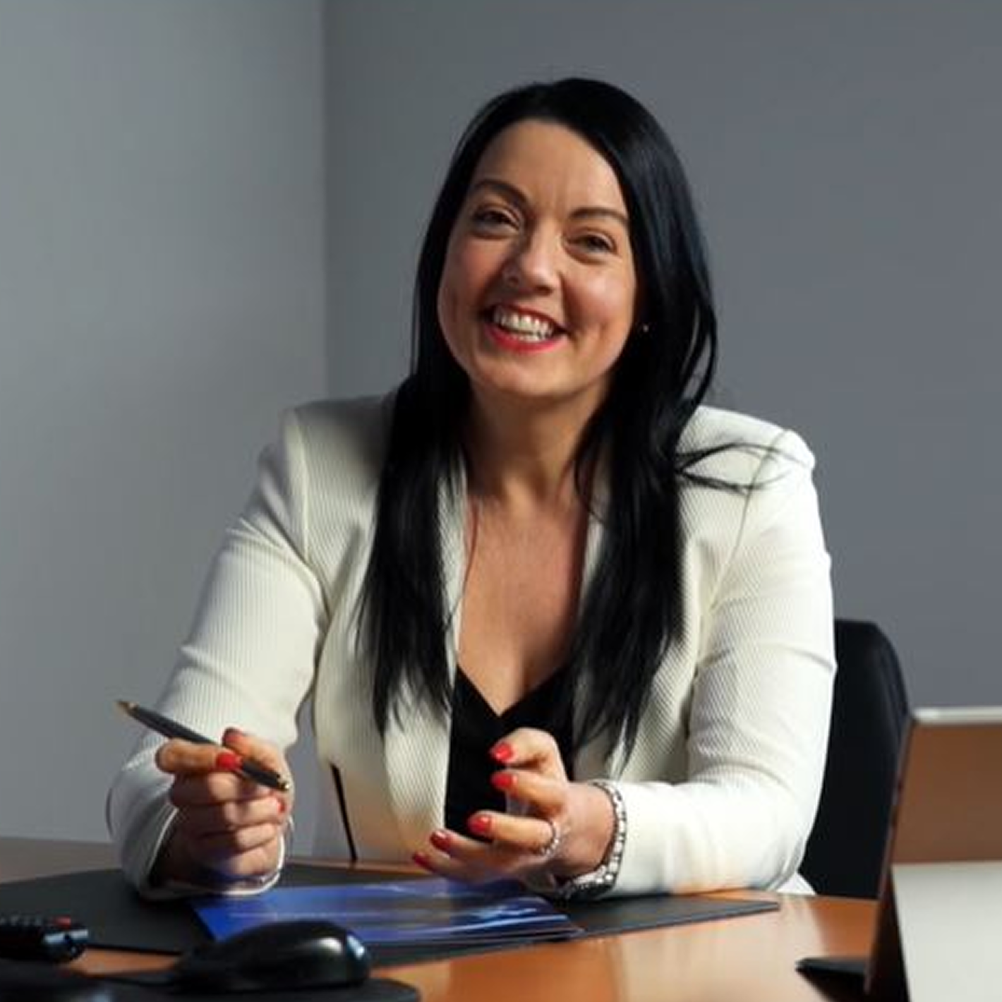 Smiling woman with dark hair in a white blazer sitting at a desk holding a pen