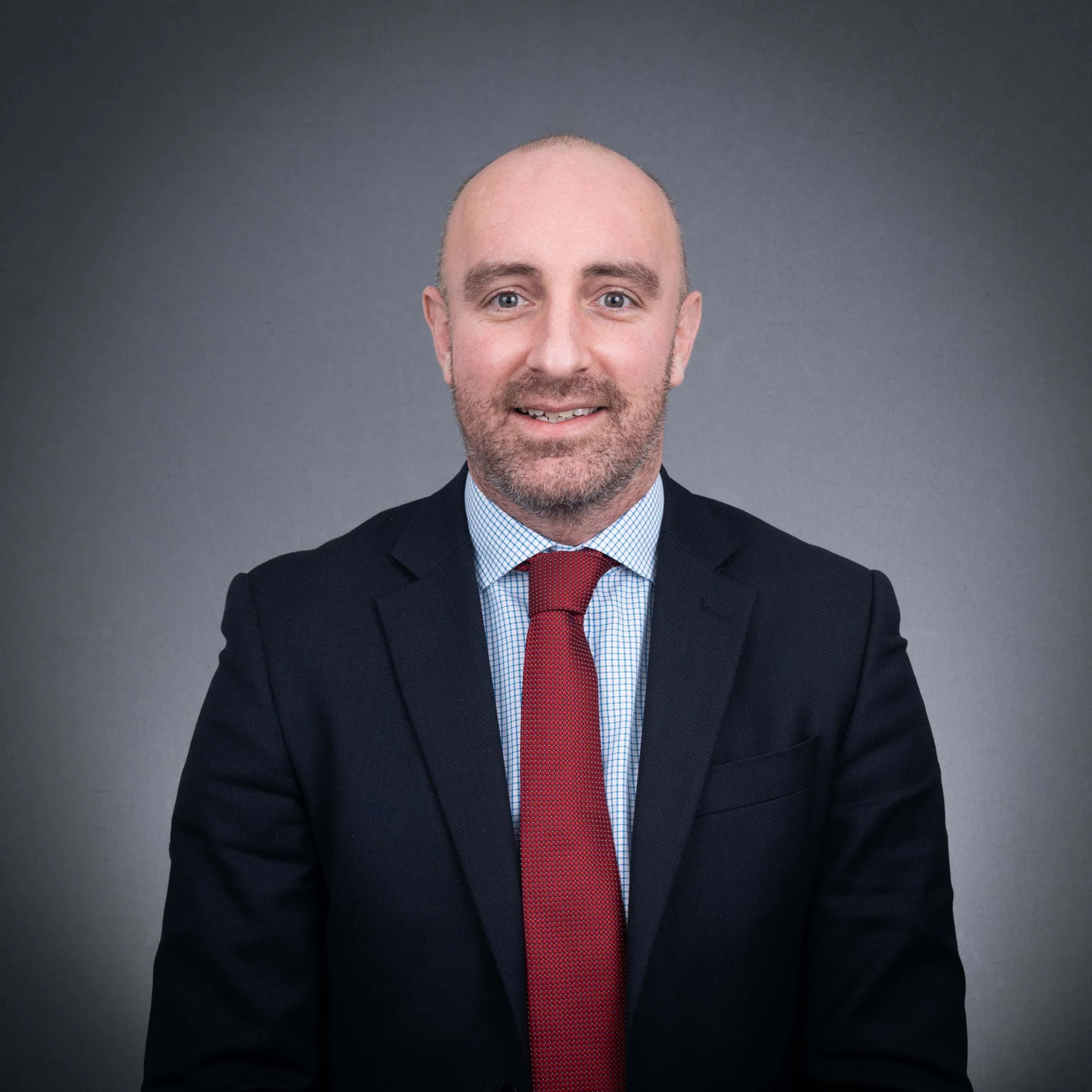 Johnny McGaughey, smiling in a dark navy suit, red tie, and checked shirt against a grey background