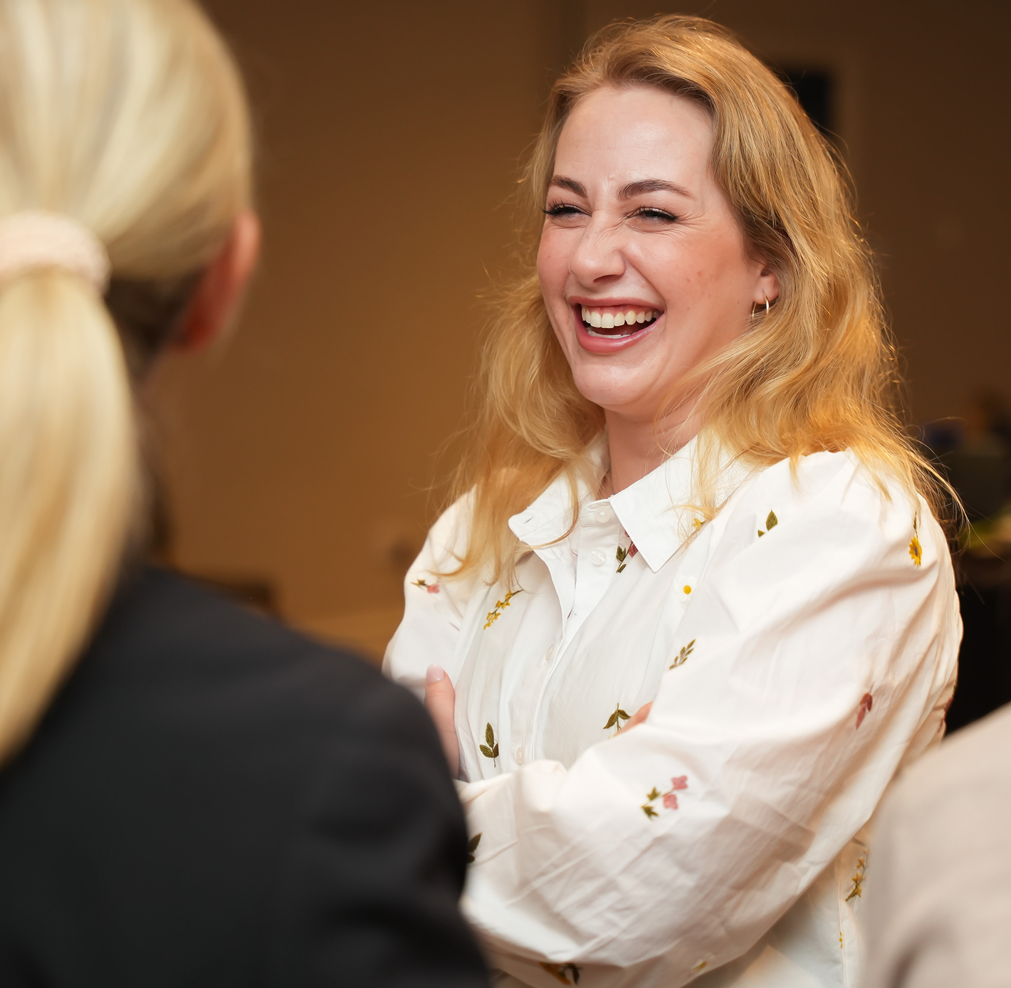 Woman in a floral white shirt laughing warmly while shaking hands with a colleague at a business meeting