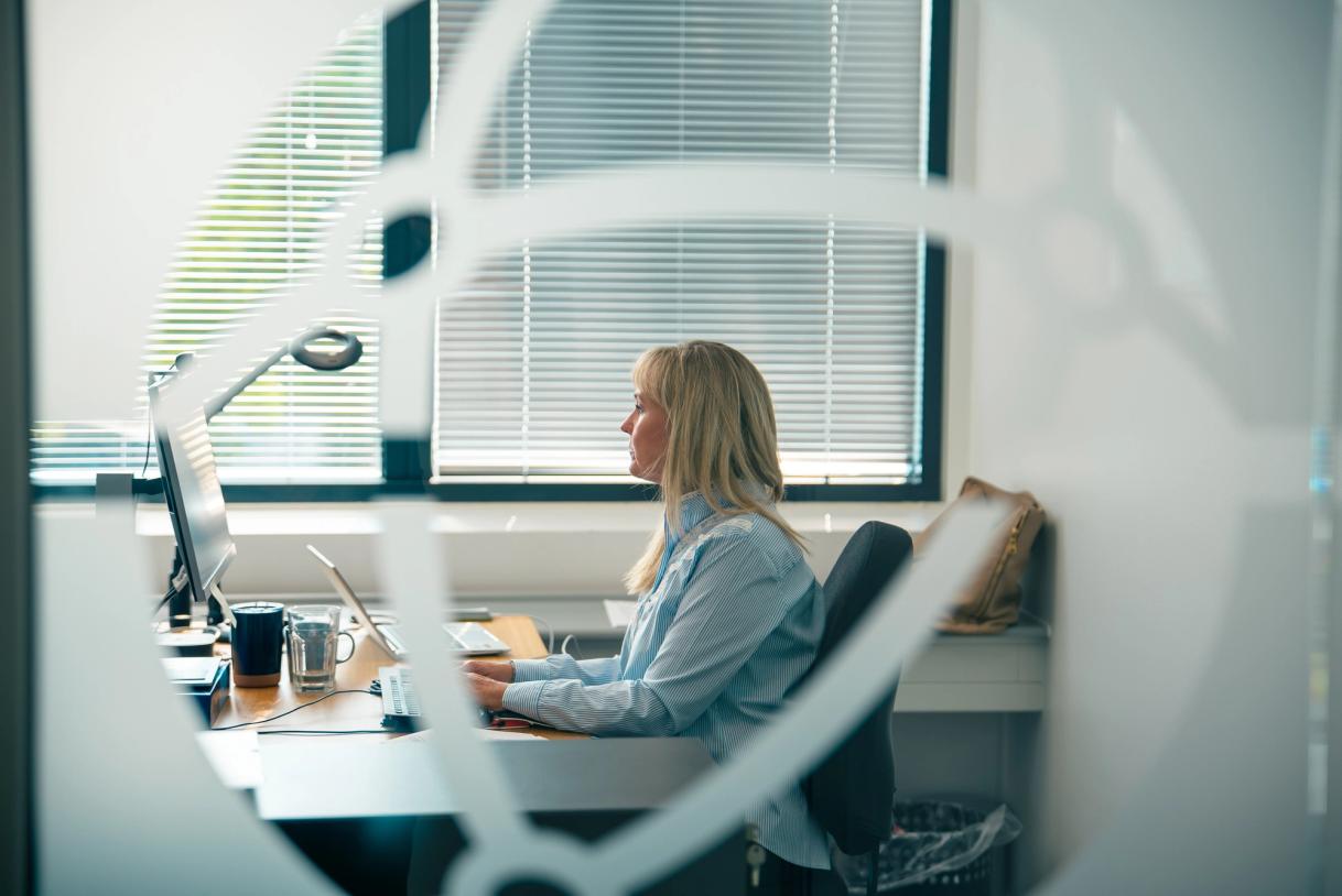 Woman working at a desktop computer in a private office, viewed through a glass partition