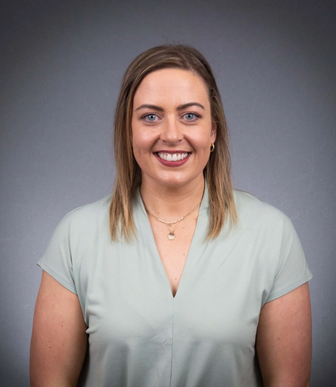 Natasha Guy smiling in a light green top and gold necklace against a grey background