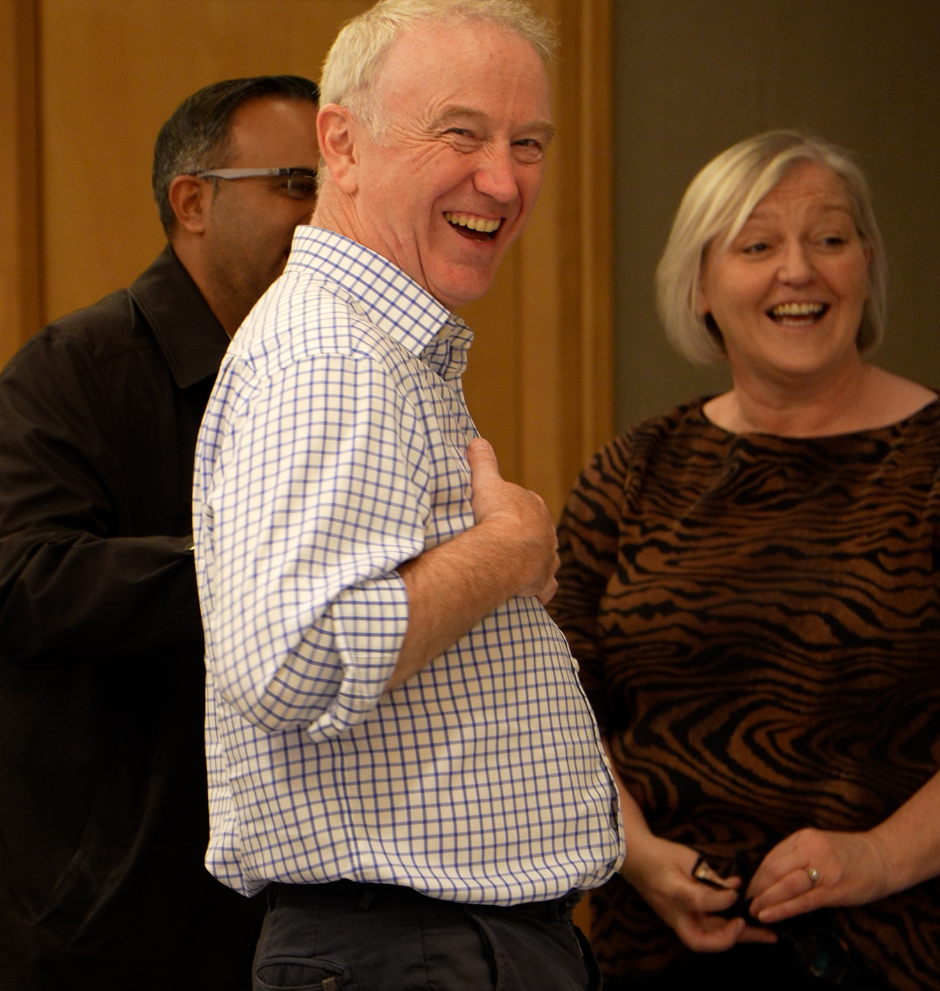 Three colleagues laughing together at a company gathering, with a man in a checked shirt prominently smiling