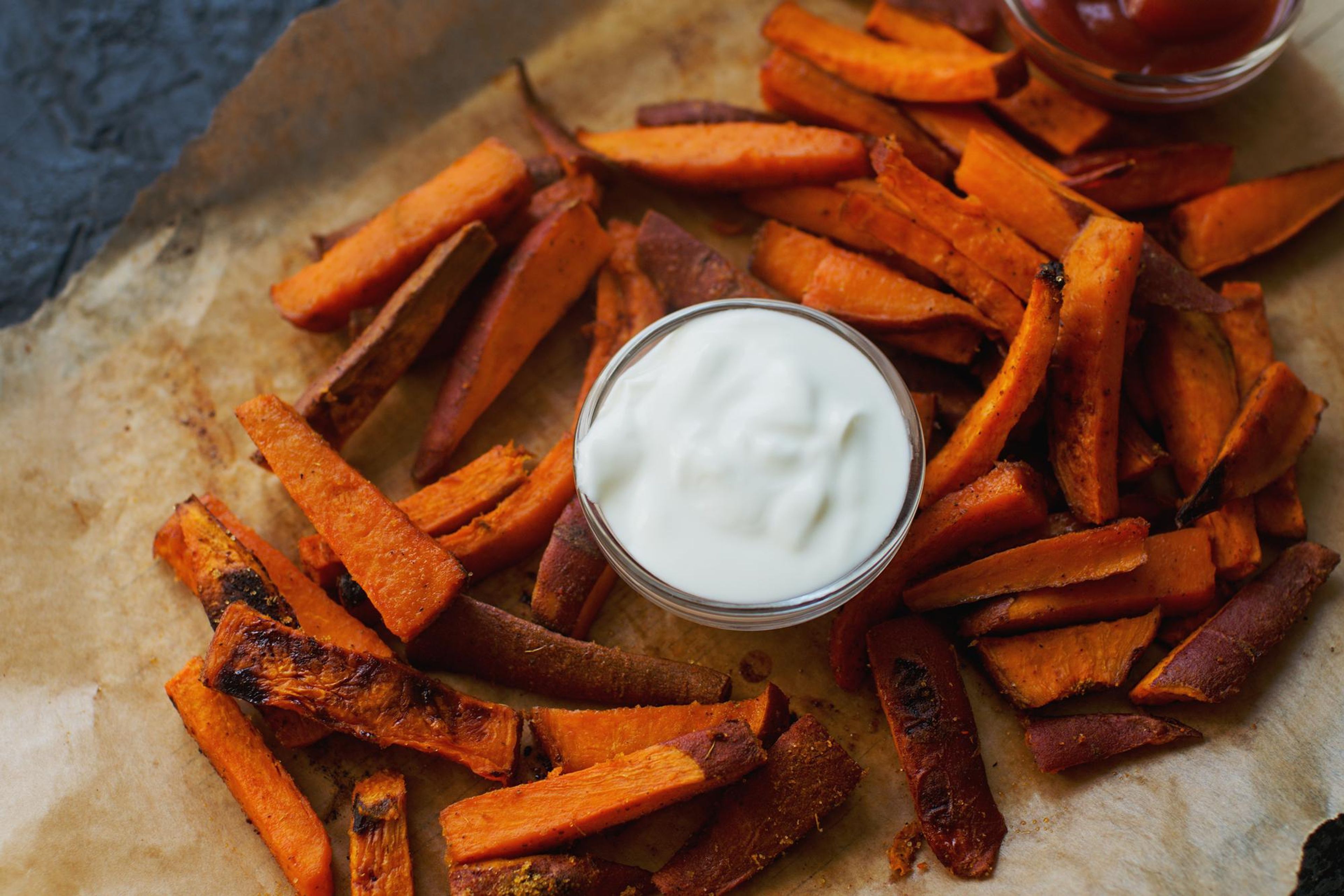 Roasted sweet potato fries with white dip and ketchup on parchment paper.