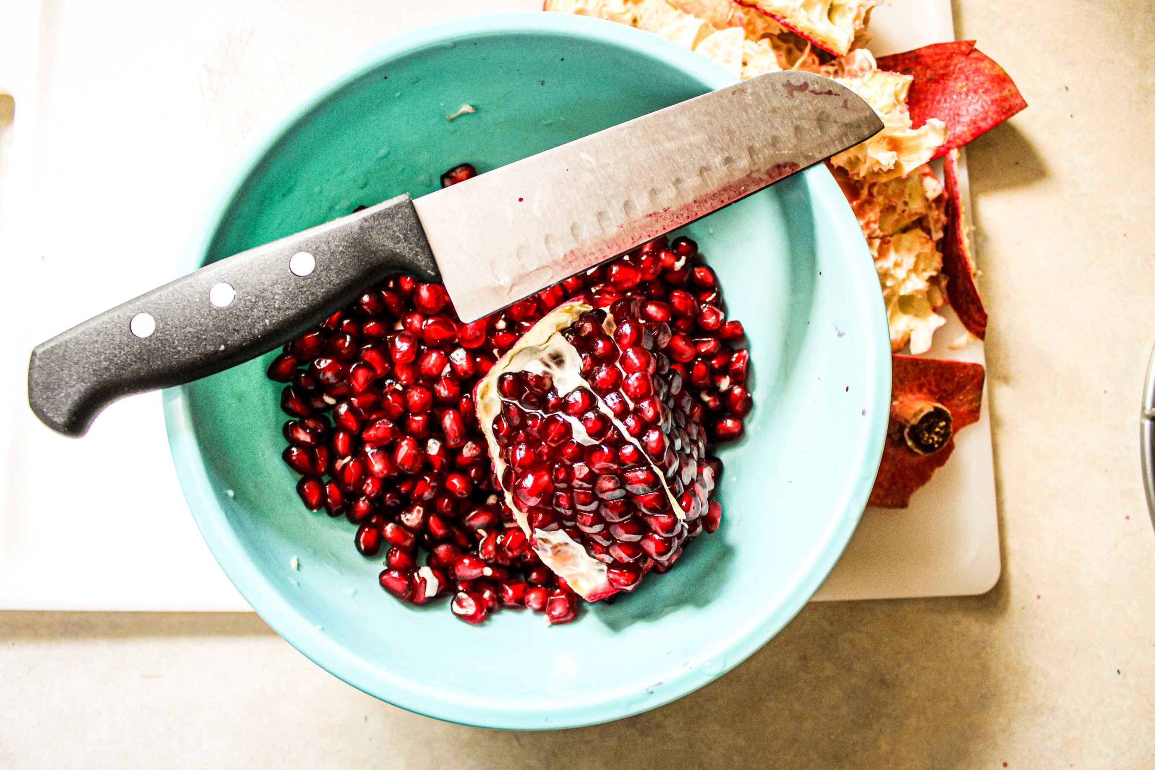A blue bowl filled with pomegranate seeds and a knife, with pomegranate peels on a cutting board beside it.