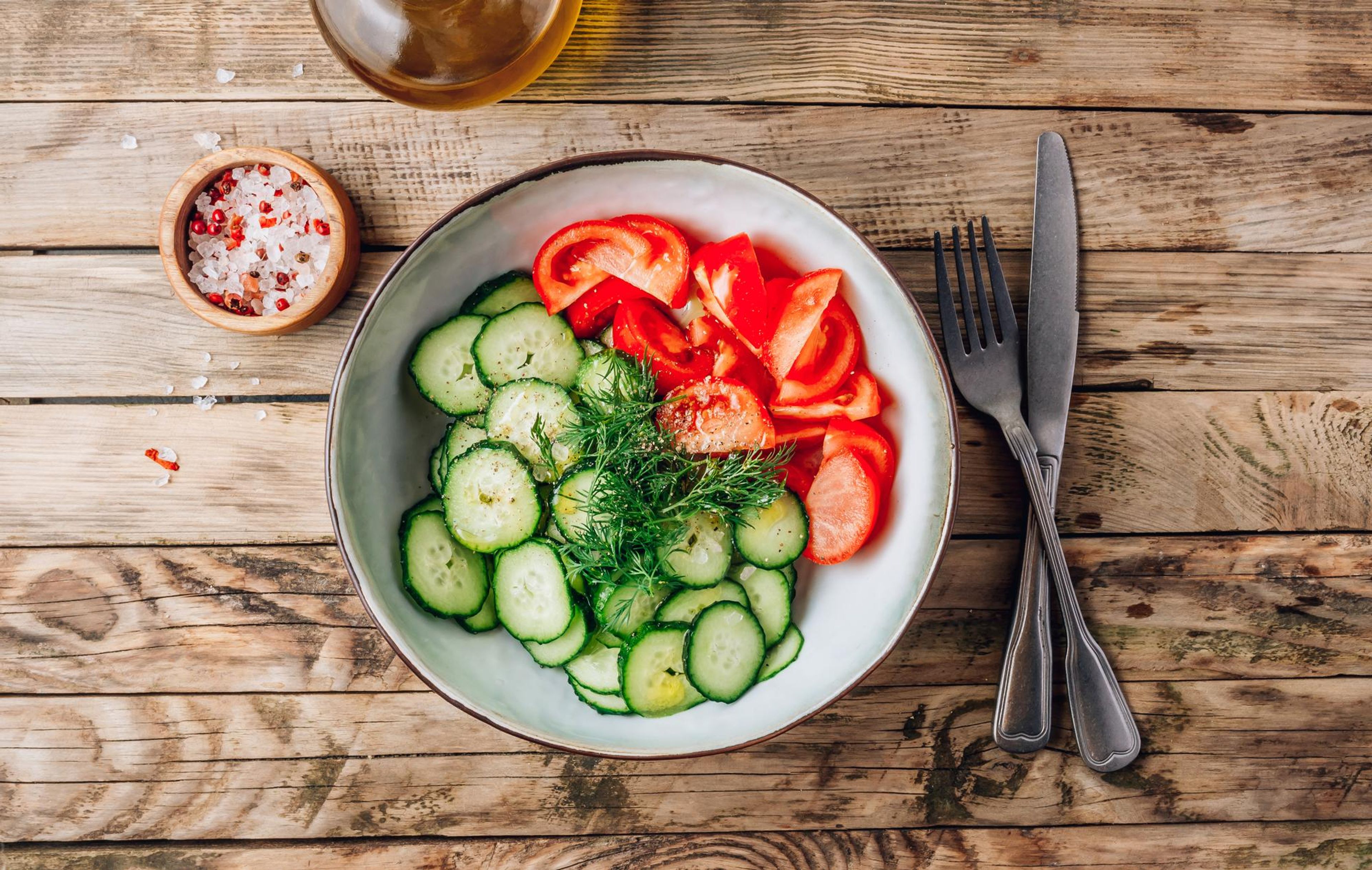 A bowl of sliced cucumbers and tomatoes with dill, on a wooden table.