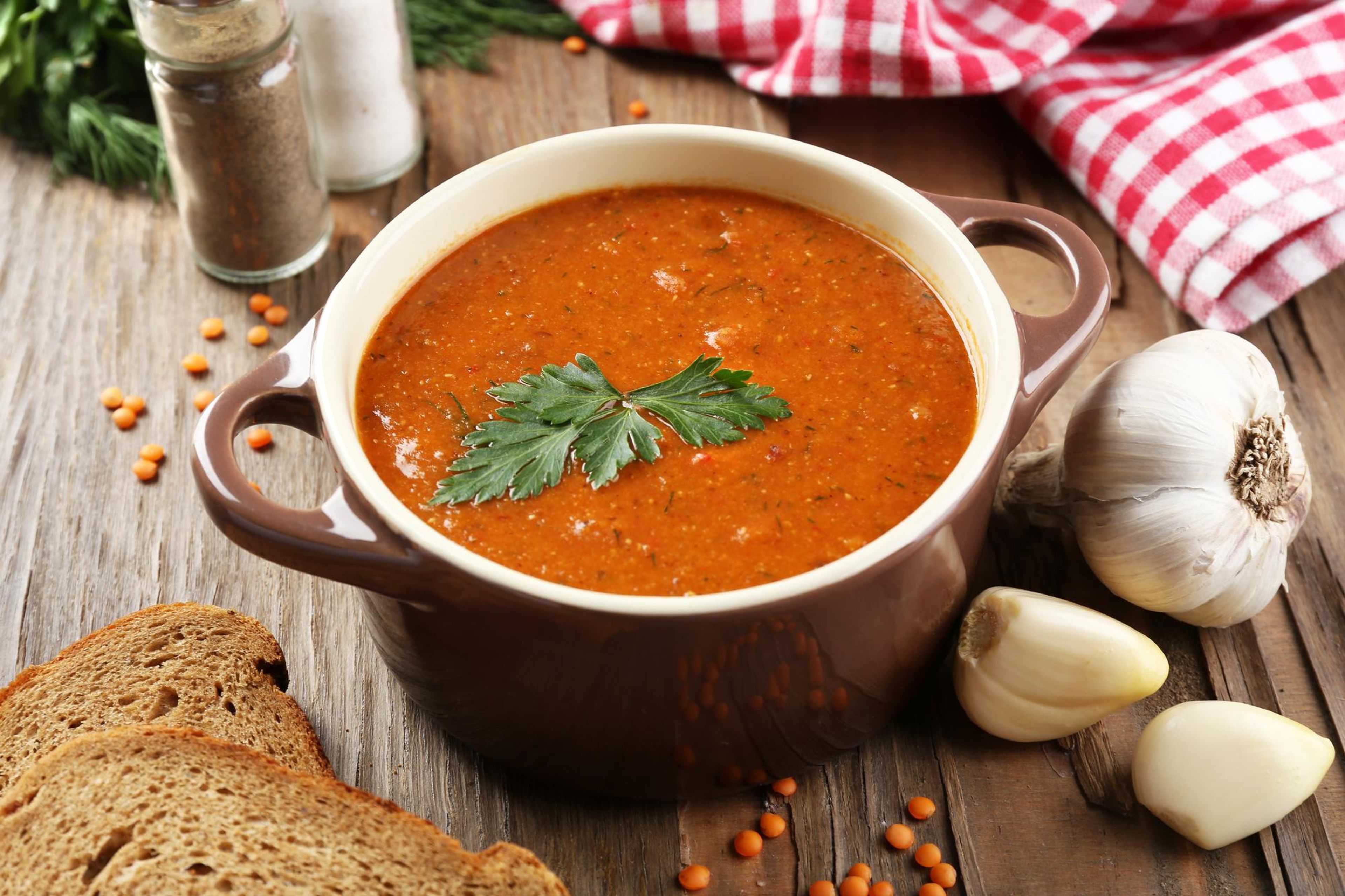 A bowl of lentil soup garnished with parsley, served with bread and garlic on a wooden table.