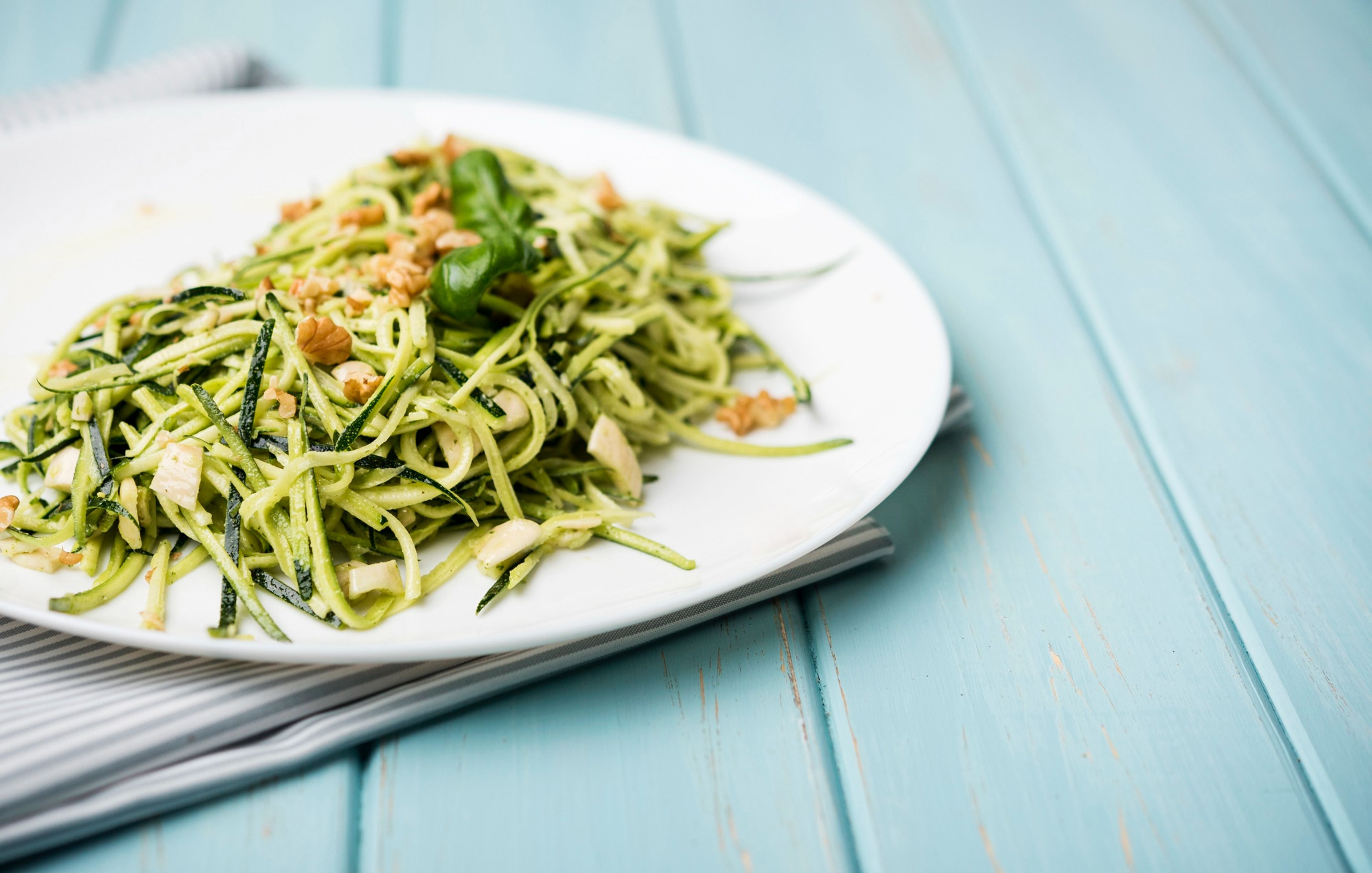 A white plate of green zucchini noodles topped with nuts and a basil leaf on a blue wooden table.