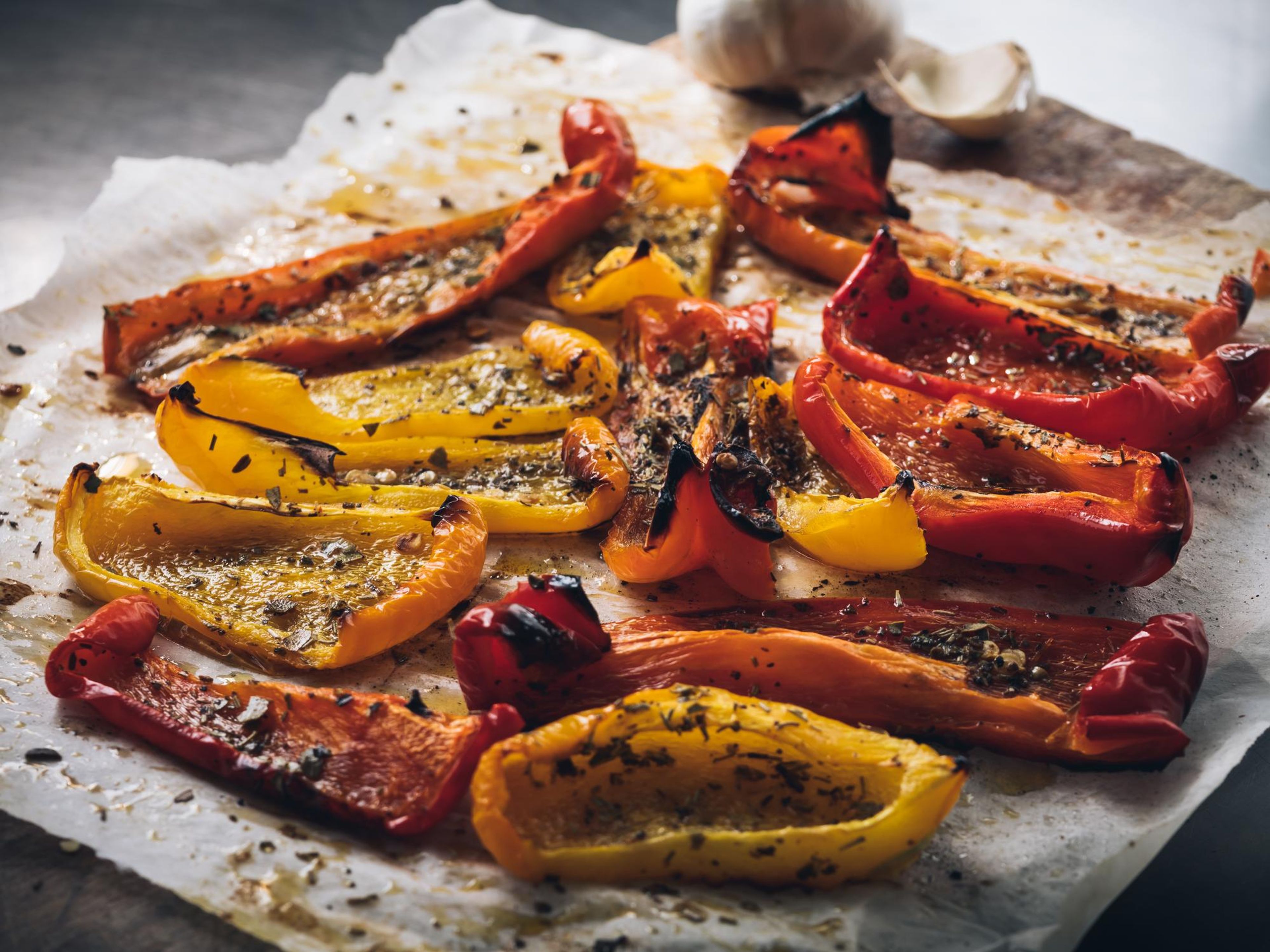 Roasted red and yellow bell peppers seasoned with herbs on parchment paper, with garlic in the background.