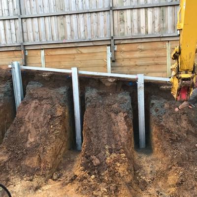 Excavator digging staged retaining wall bays below an existing fence, leaving soil piers to protect the neighbouring property