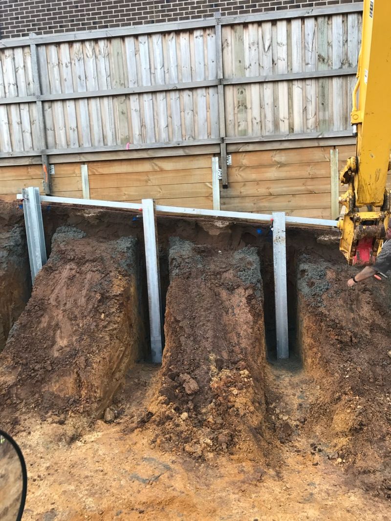 Excavator digging alternating staged bays below an existing boundary fence
