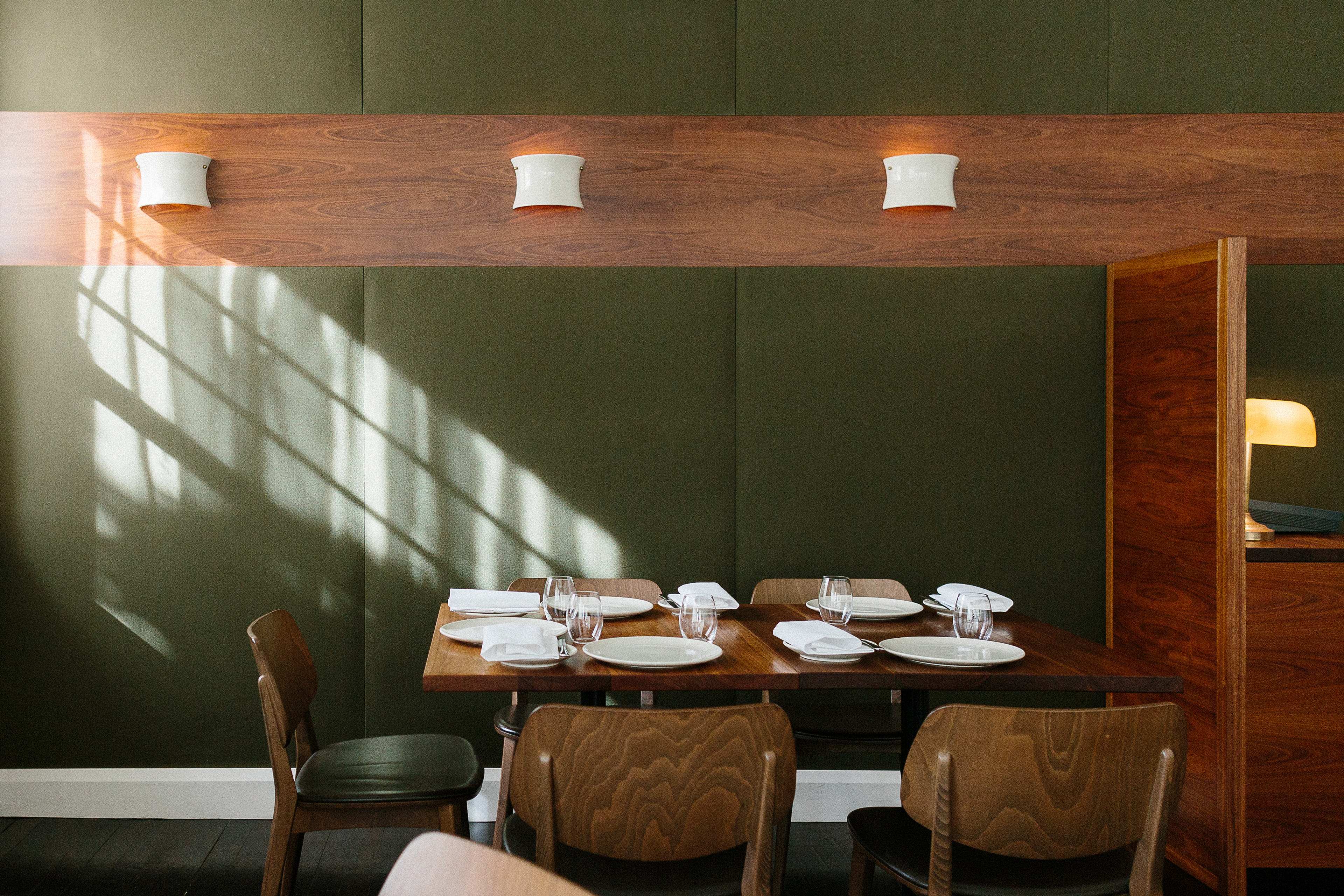 A solid timber dining table and chairs sits in front of green upholstered walls and custom ceramic wall lights at Sixpenny, a locals' favourite restaurant in Sydney - designed by Foolscap.