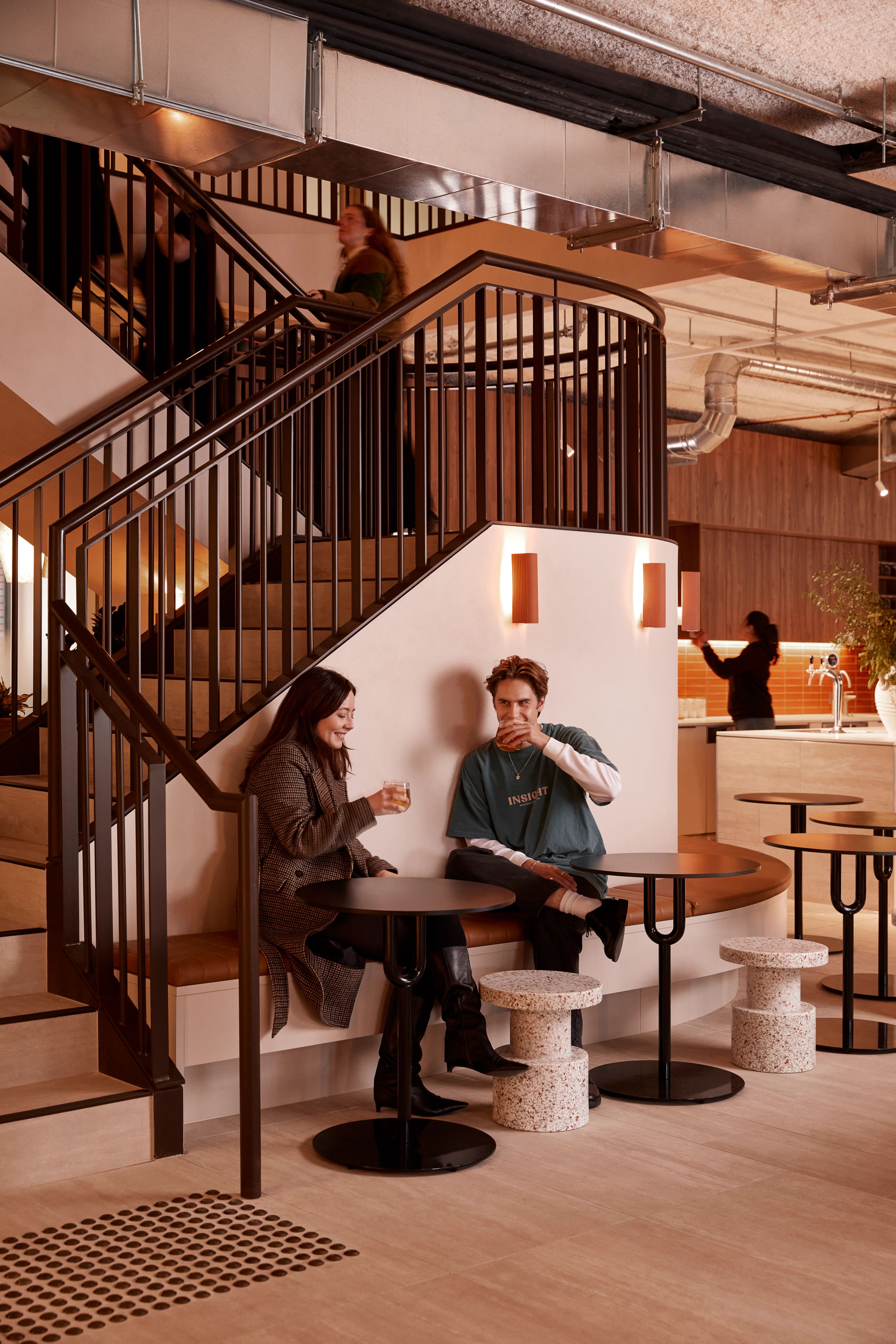 Two people sharing a drink on a booth seat which circles a connecting stair at Forum Workspace, a co-working space designed by Foolscap. 